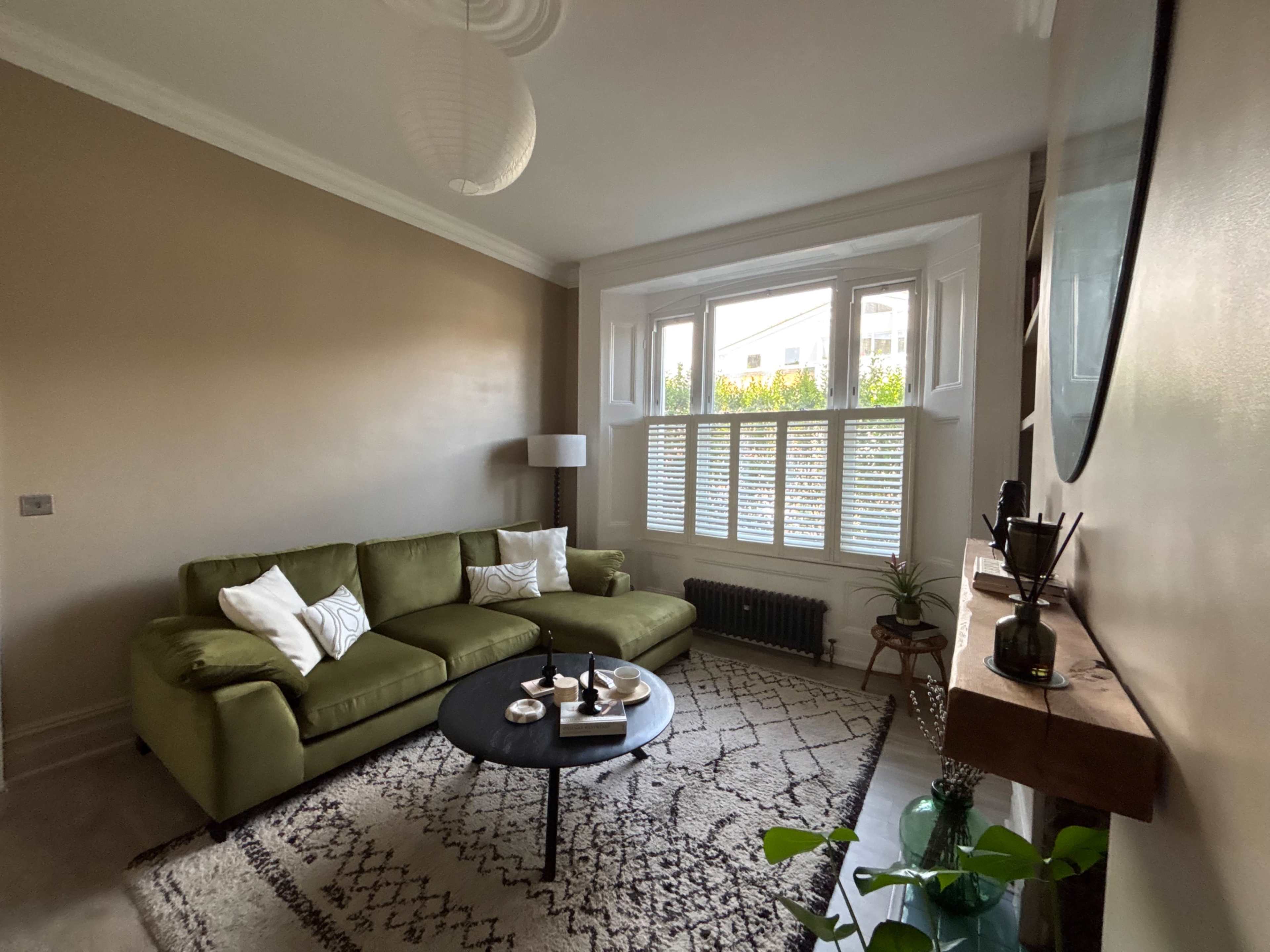 The living room features a green sectional sofa, a round black coffee table, a lamp, and a window with plantation shutters.