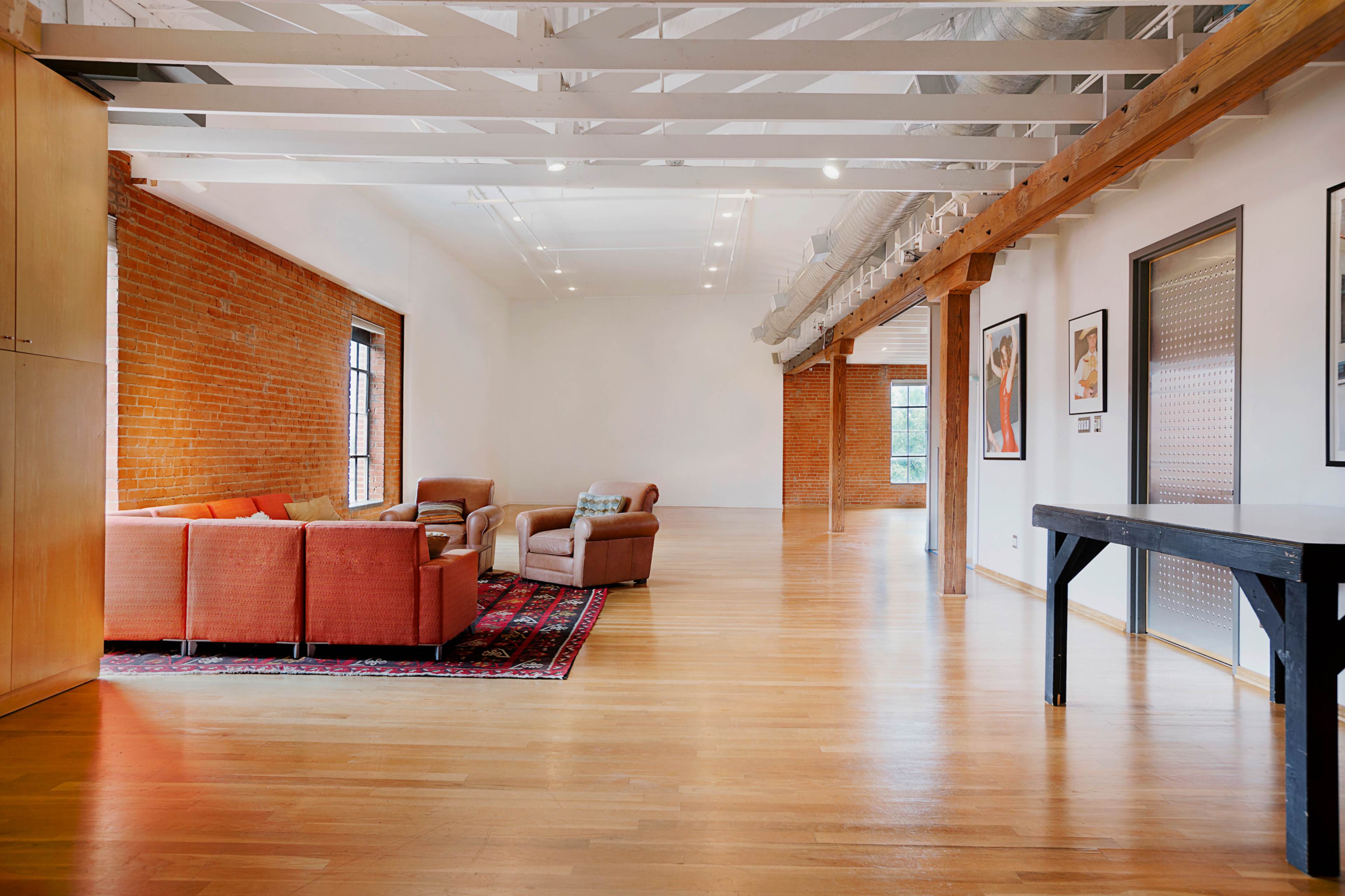 A spacious living area with exposed brick walls, wooden beams, and polished hardwood floors, featuring a sofa set and a dining table.