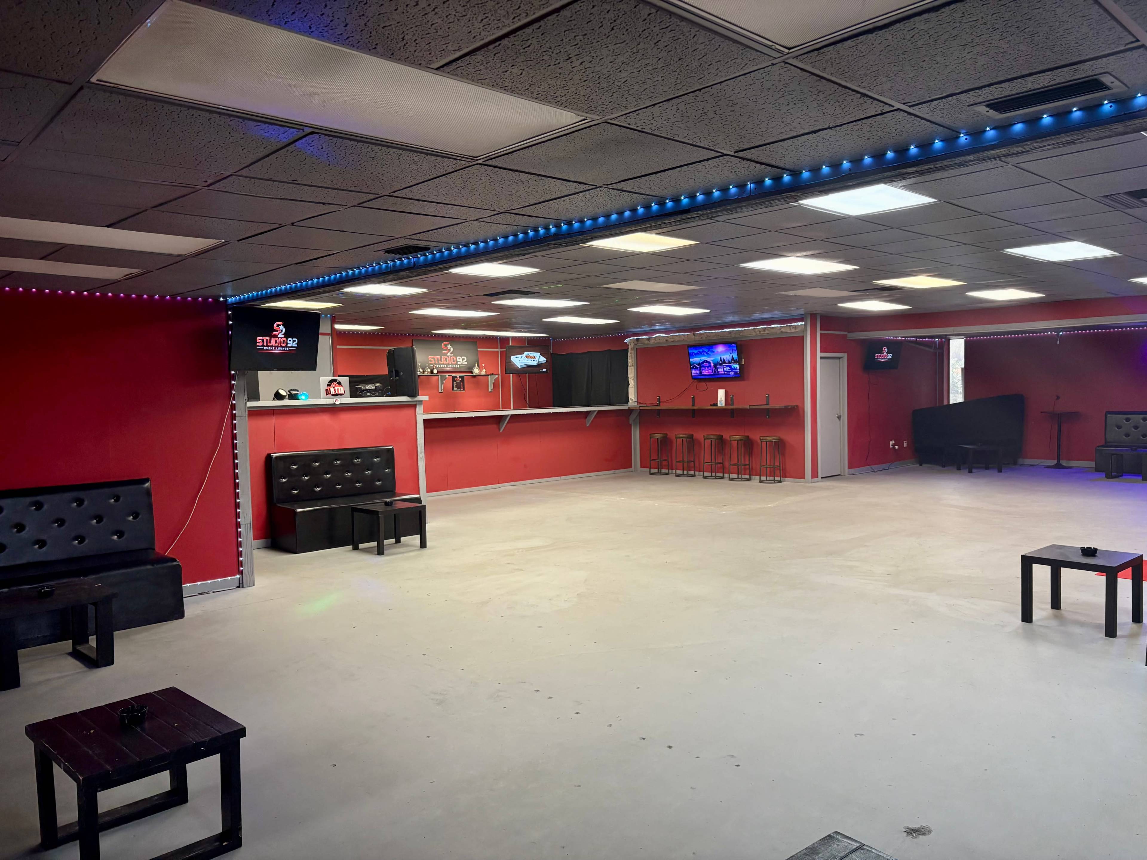 The image shows a spacious, empty room with red walls, black furniture, and a bar area featuring stools and a television display.