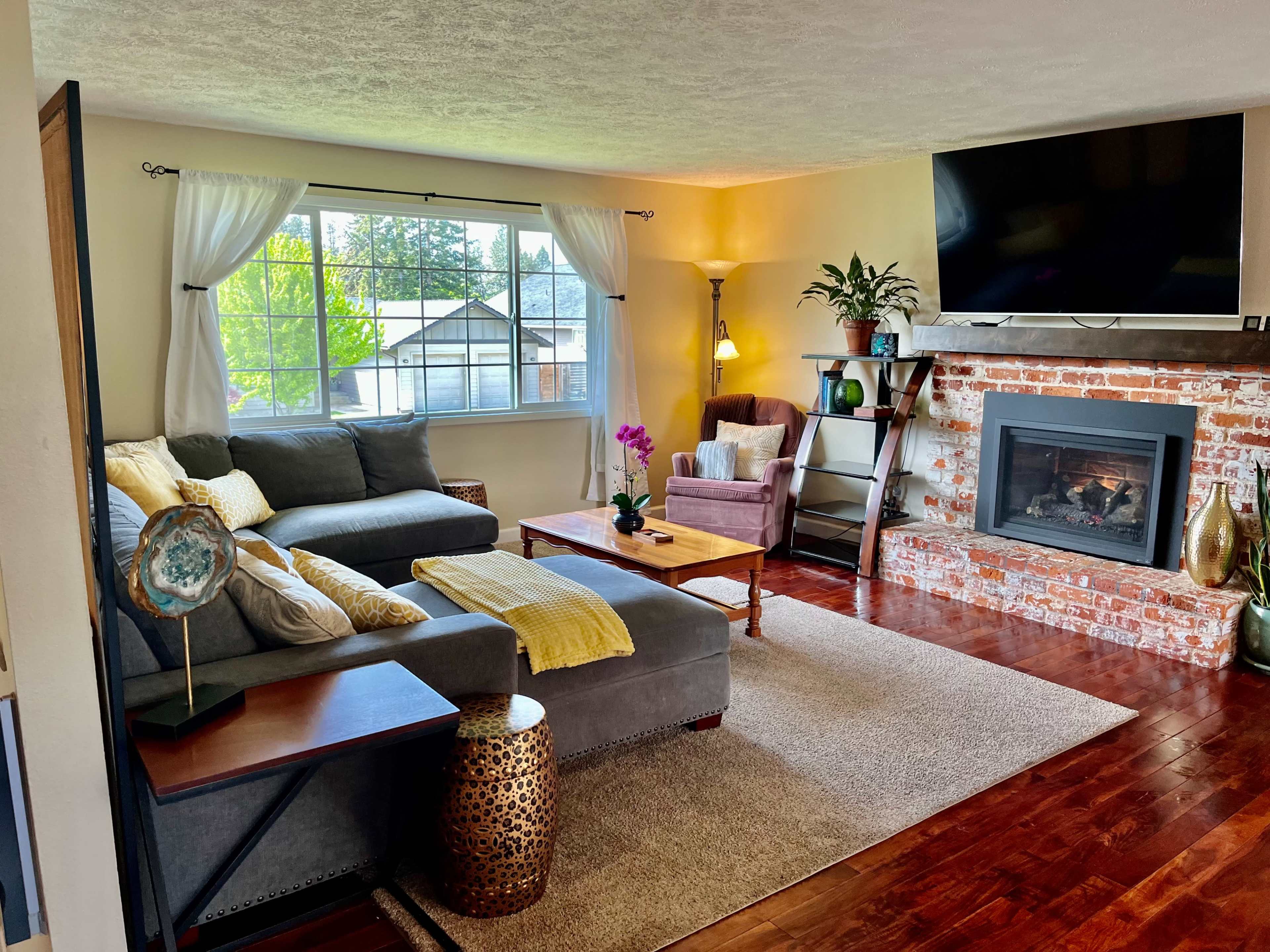 The living room features a gray sectional sofa, a brick fireplace, and a large window with natural light illuminating the space.