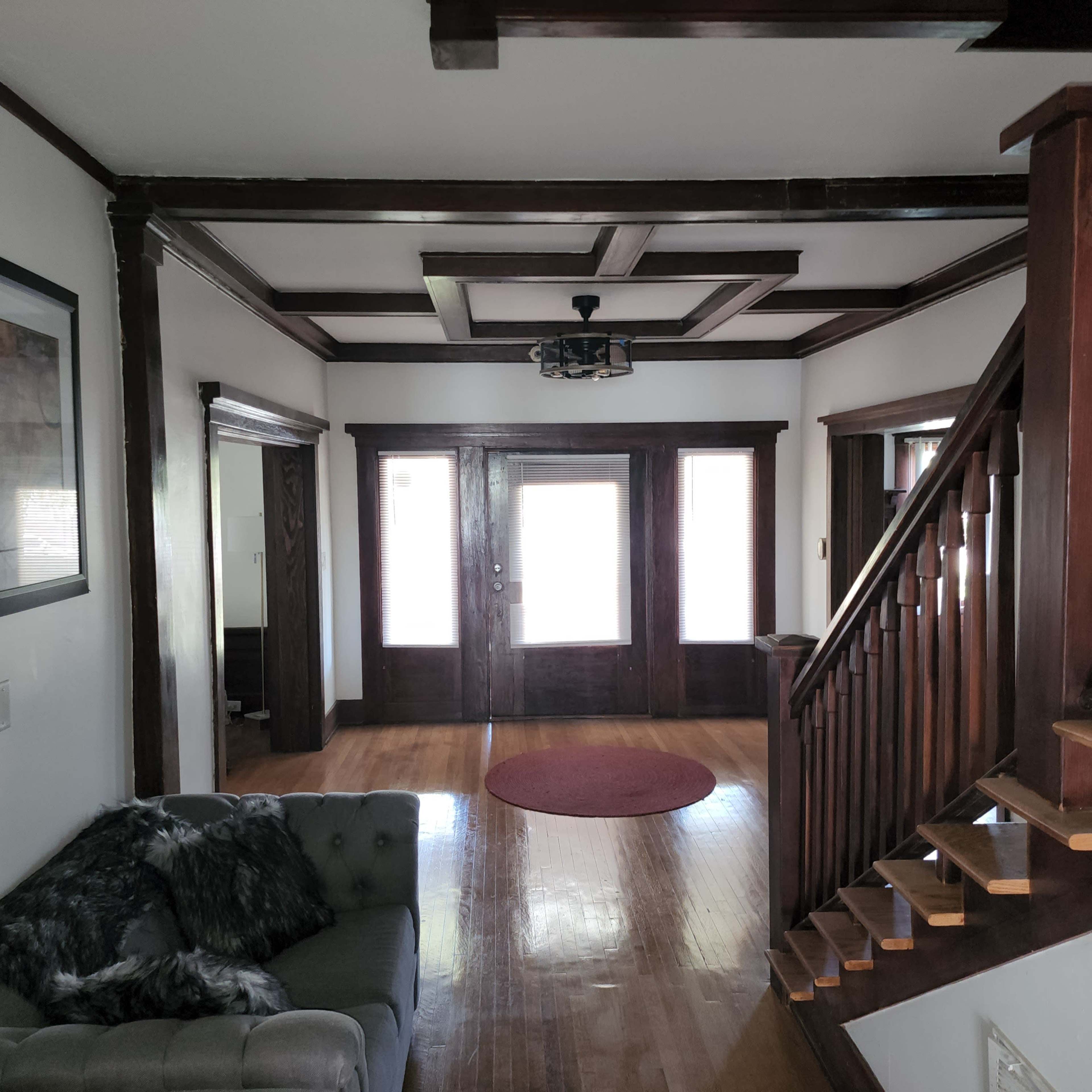 The image shows a house entrance with a staircase on the right, a circular rug in the center, and large windows allowing light to enter.