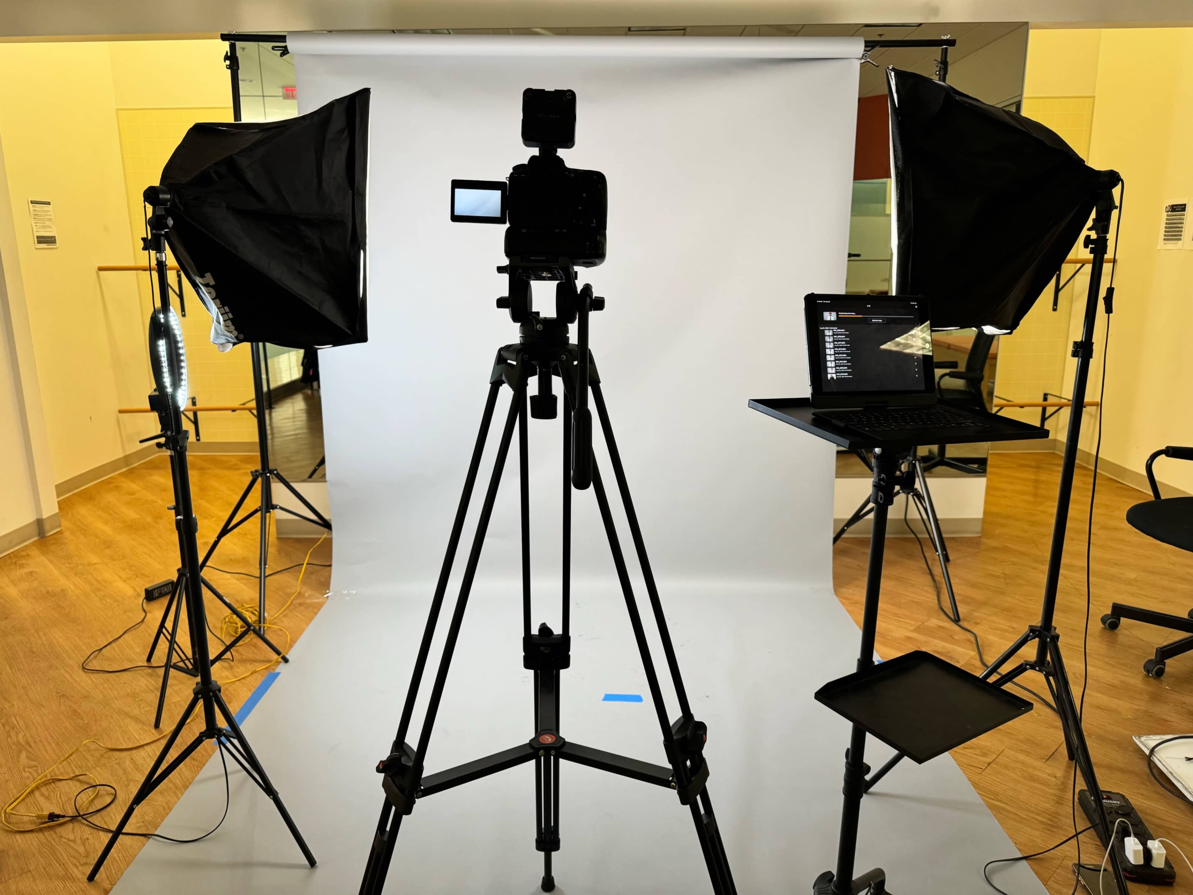 The image shows a photography studio setup with a camera mounted on a tripod, flanked by two softbox lights and a laptop on a stand, all positioned in front of a white backdrop.