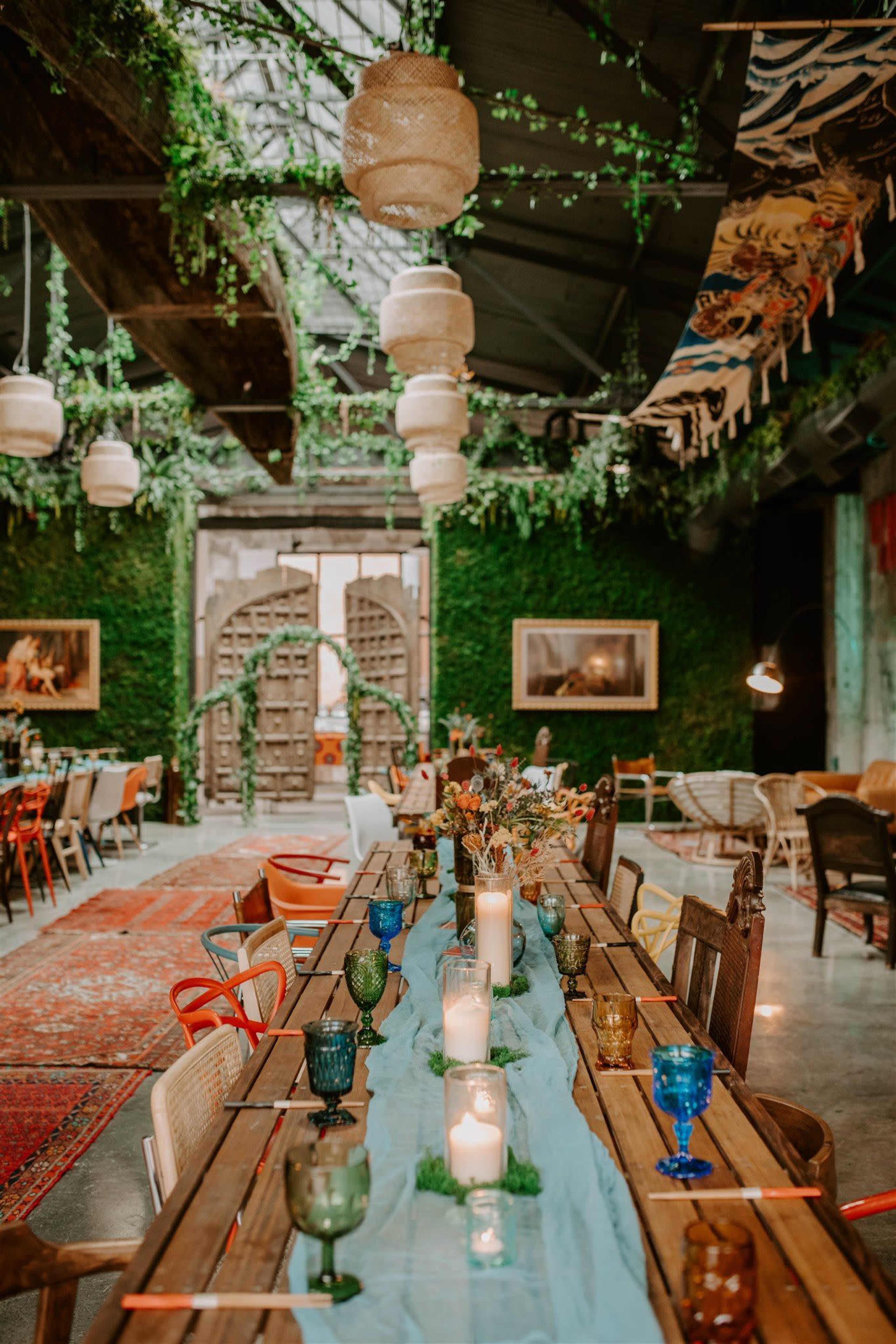A long wooden dining table is set with colorful glassware and candles, surrounded by greenery and decorative lighting in a spacious indoor venue.
