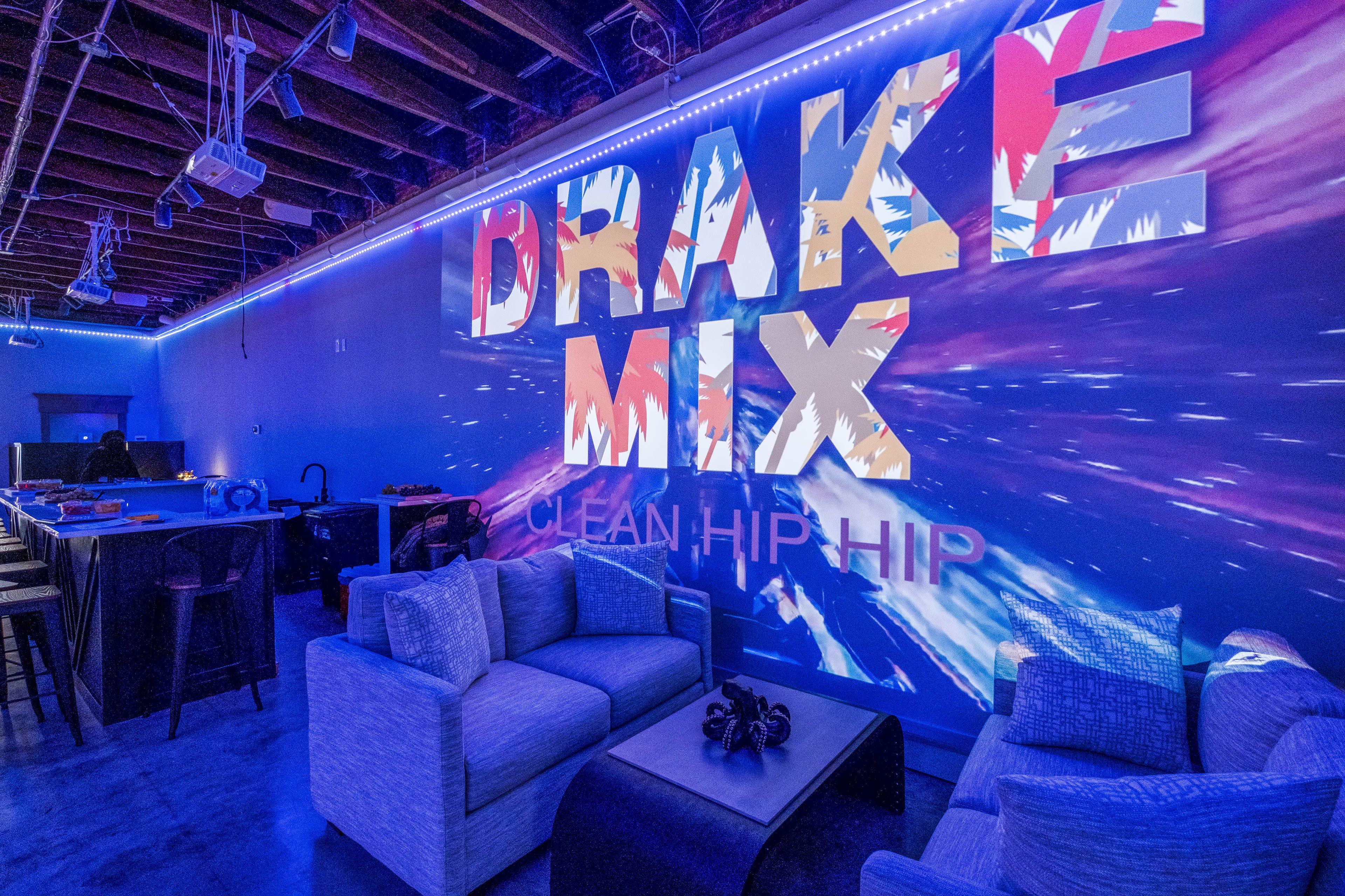 A modern lounge features a large wall display advertising "DRAKE MIX" with colorful graphics, surrounded by stylish seating and a bar area.