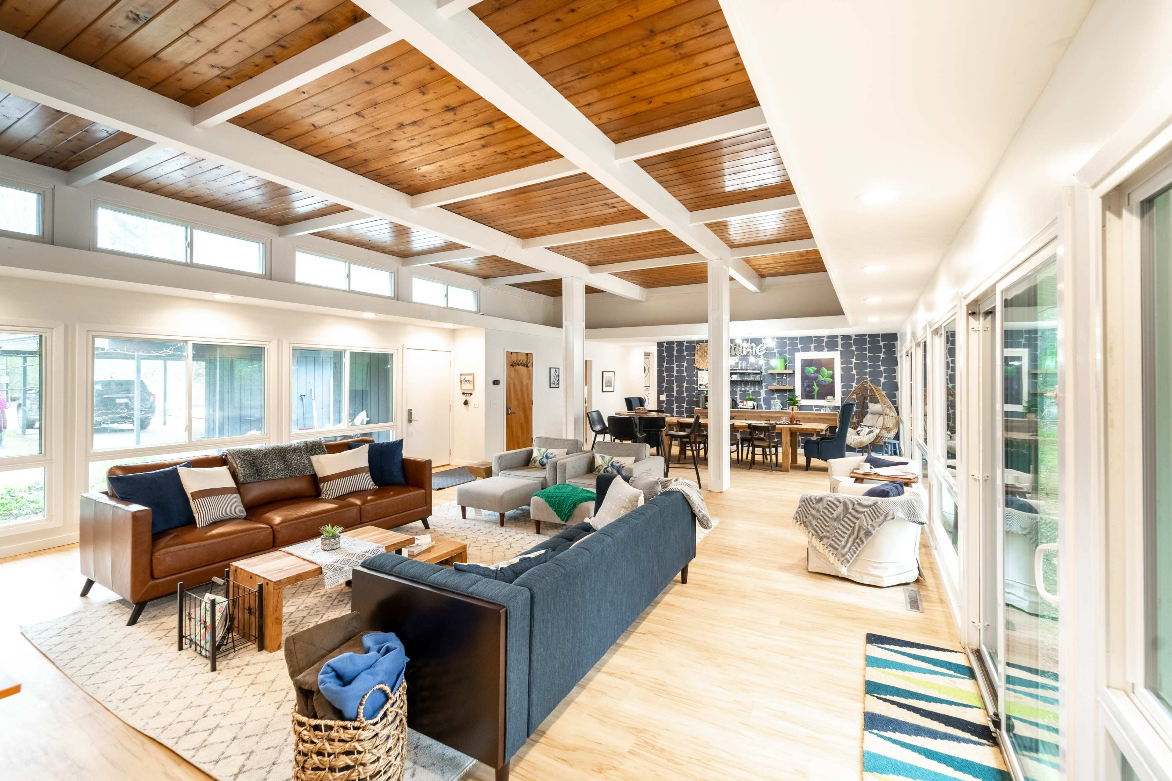 A spacious living area with a mix of leather and fabric sofas, a wooden ceiling, large windows, and a dining table in the background.