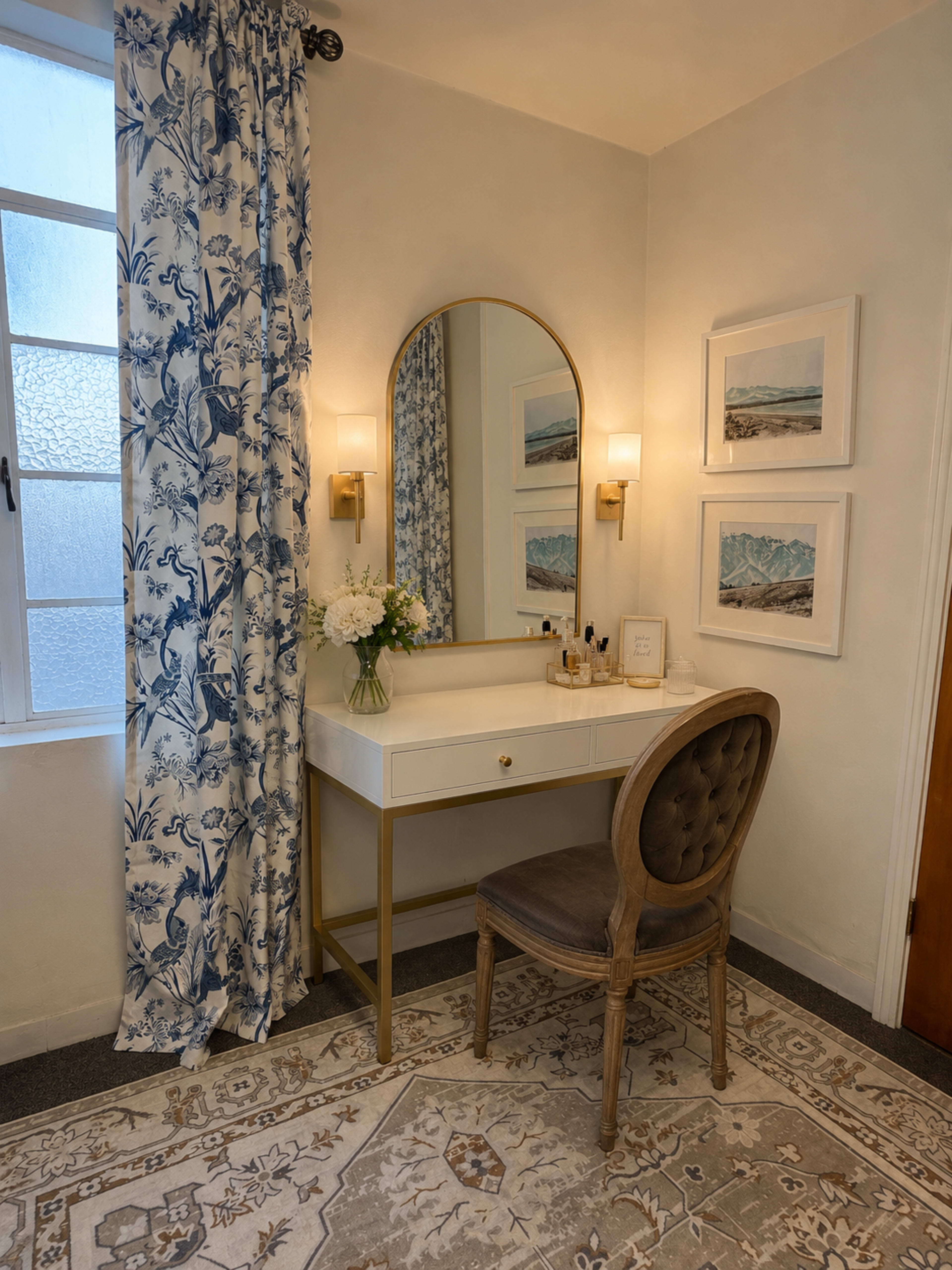 The image shows a styled vanity area featuring a round mirror, a decorative chair, and a flower vase in a softly lit room with floral curtains and framed artwork on the walls.