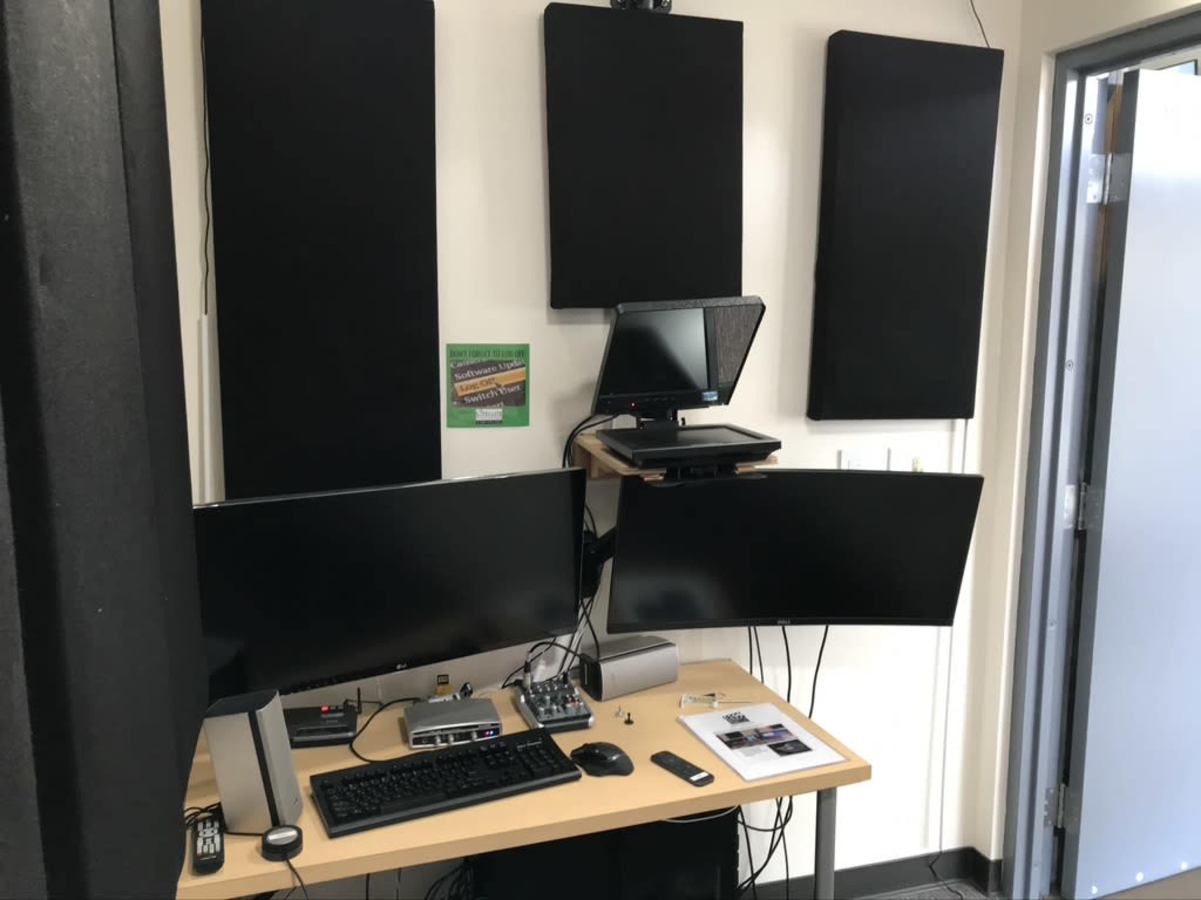 The image shows a small workspace with two computer monitors, audio equipment, and soundproofing panels on the wall.