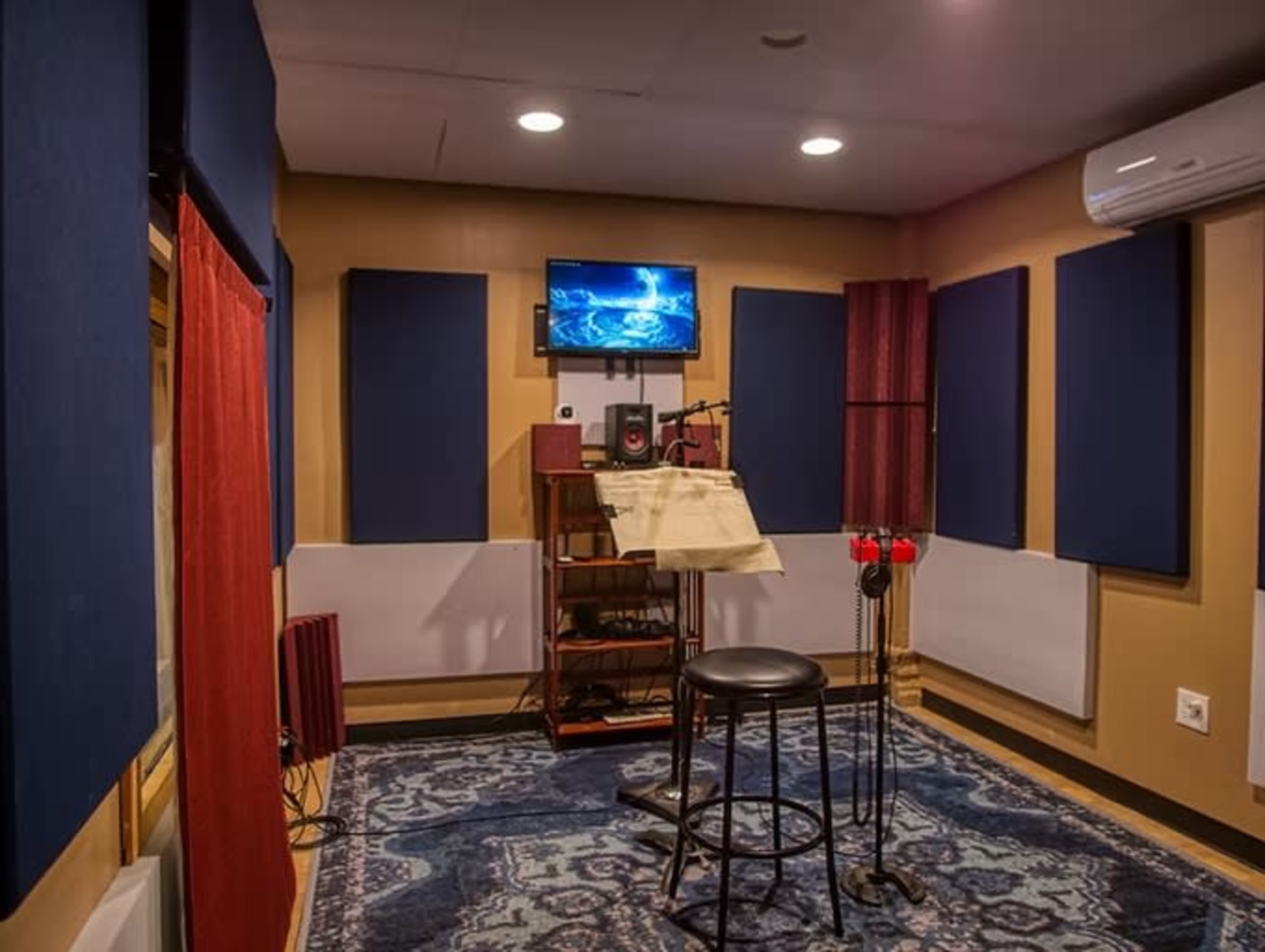 The image shows a small recording studio with a microphone and a music stand in the center, surrounded by soundproof walls and equipped with a monitor and audio equipment.