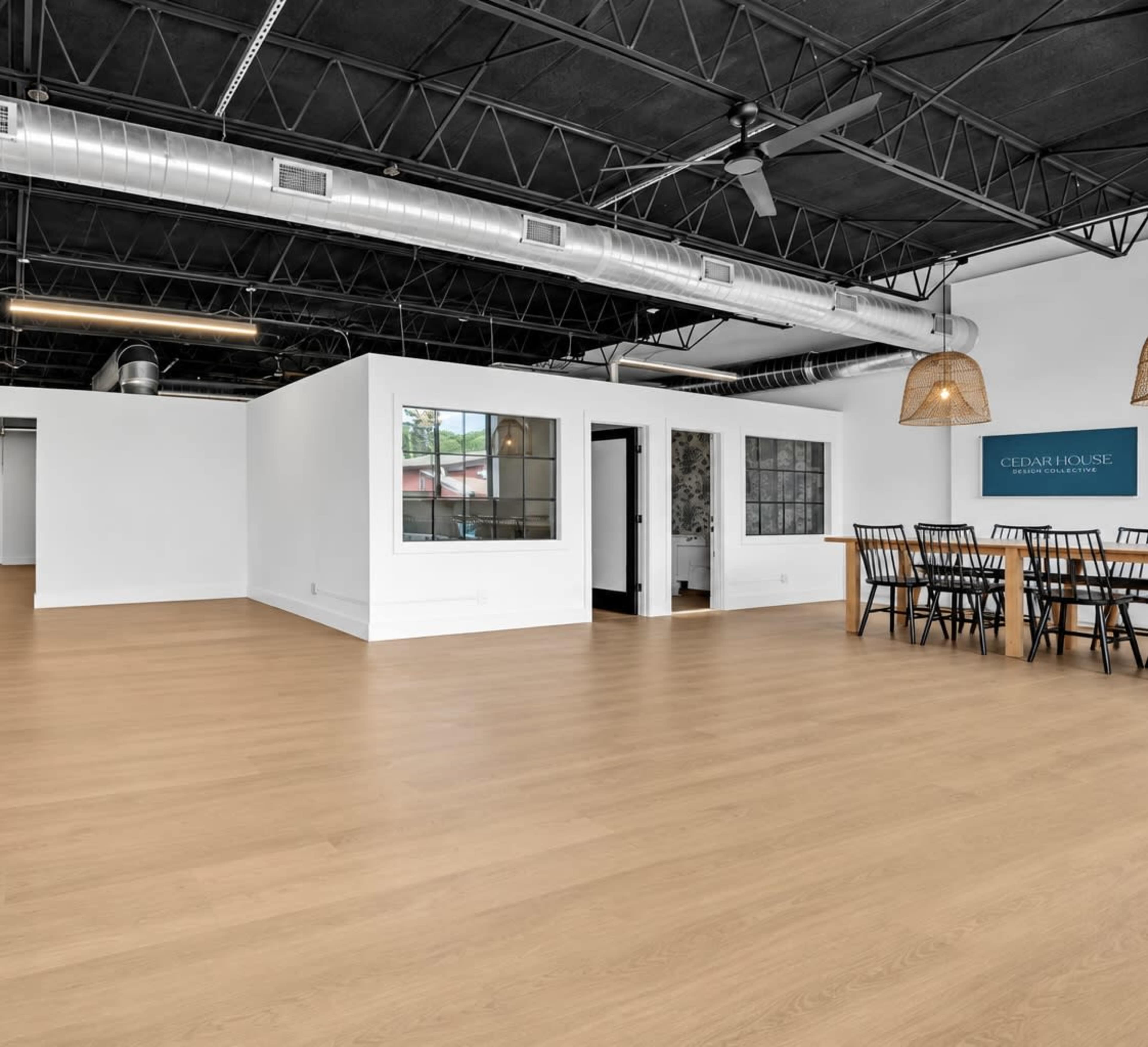 The image shows a spacious, open room with light wood flooring, featuring a dining table with chairs, a small enclosed area with windows, and a blue wall with the text "CEDAR HOUSE."