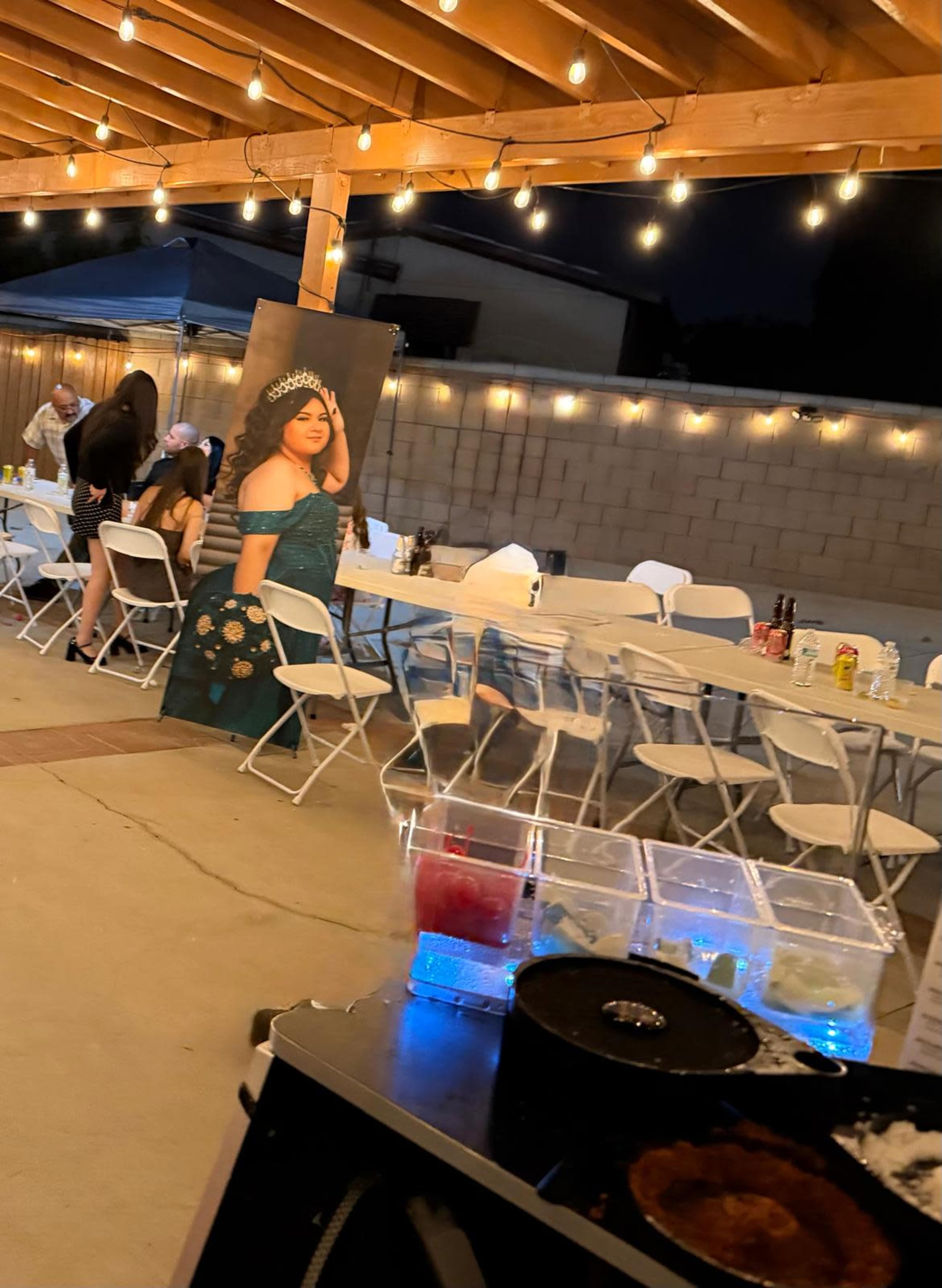 The scene features a decorated outdoor space with tables and chairs under string lights, accompanied by a large poster of a woman in a dress.
