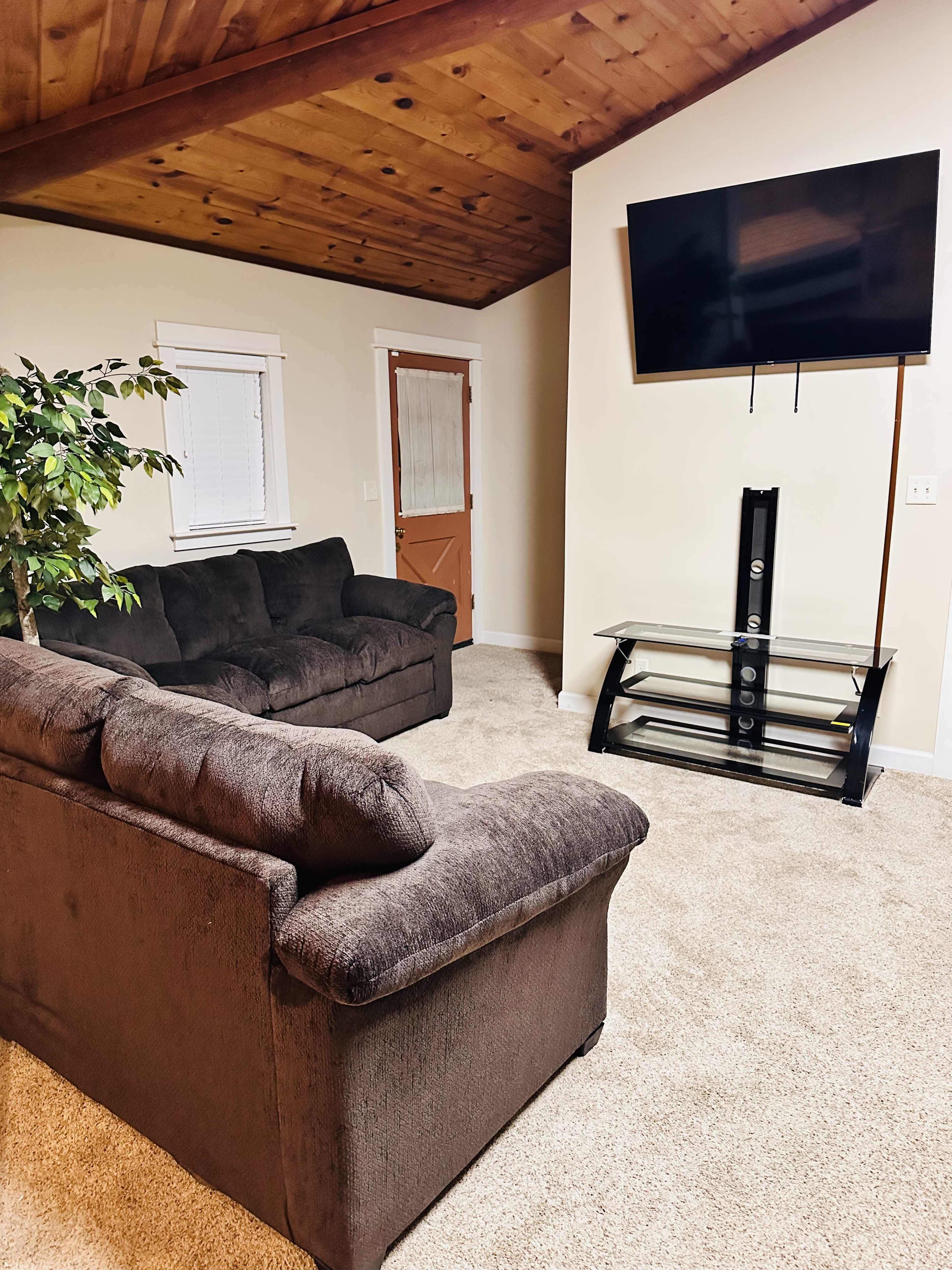 A living room with a dark couch, a glass TV stand, and a mounted television on a wall with a wooden ceiling.