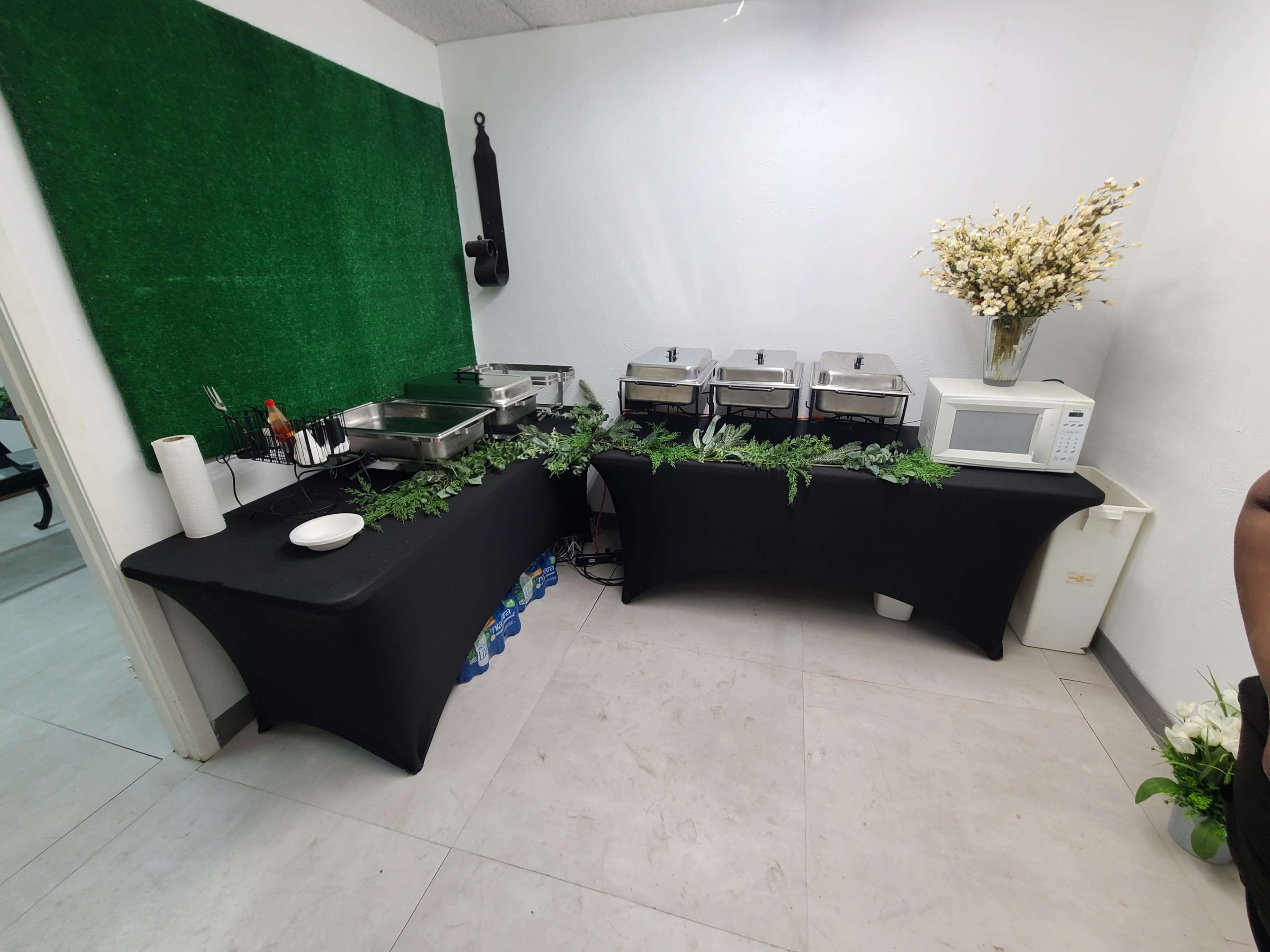 A buffet setup with several food warmers, a microwave, and decorative greenery arranged on a black tablecloth against a green backdrop.