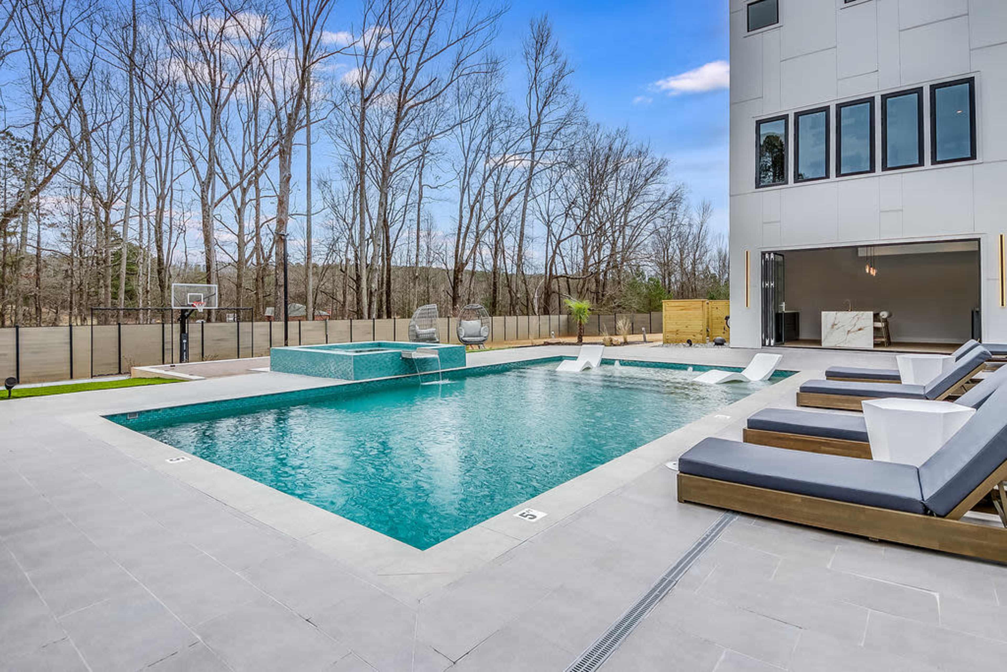 A modern swimming pool surrounded by loungers and a basketball hoop in a wooded setting.