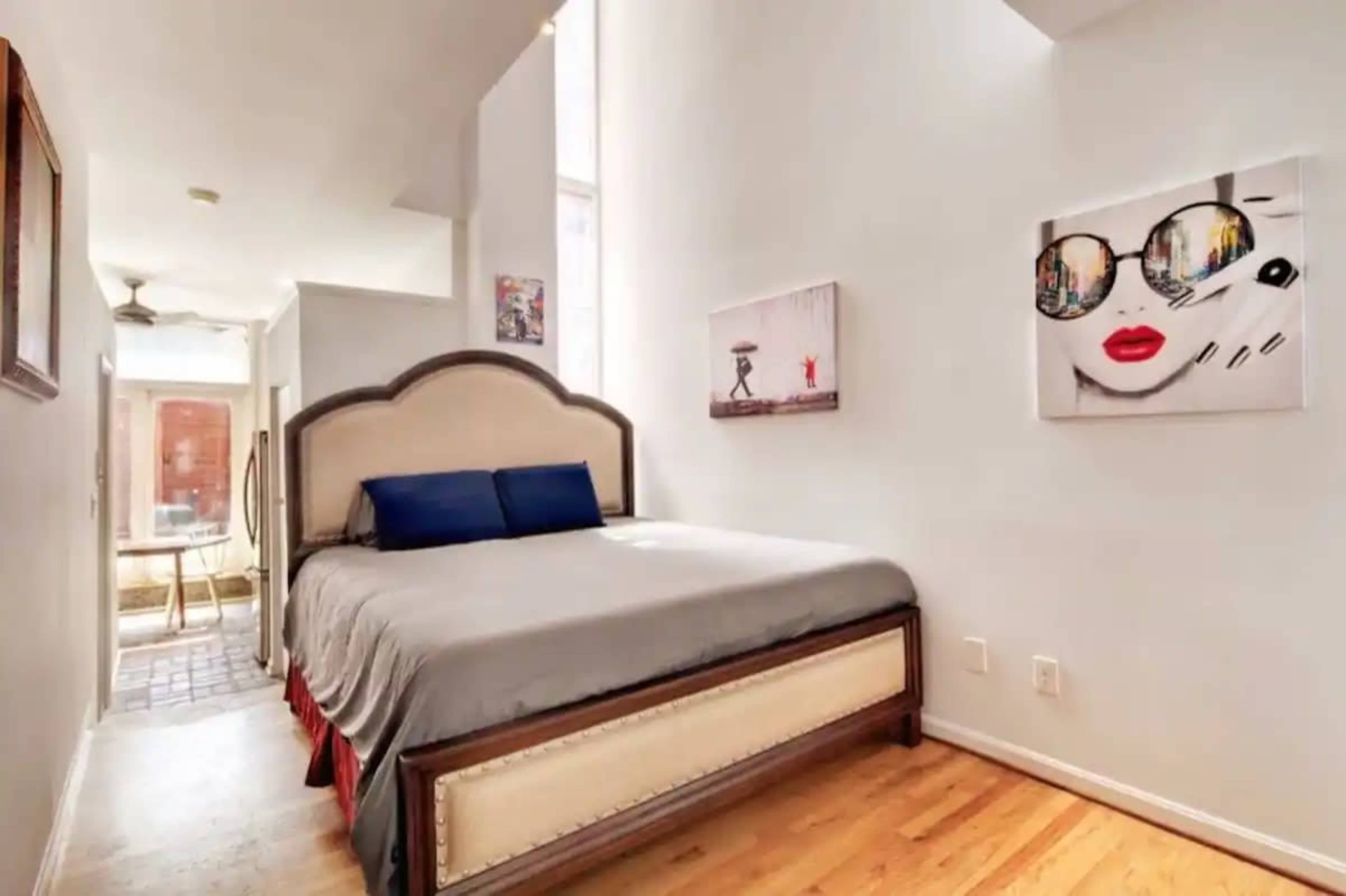 A bedroom with a large bed, two blue pillows, and artwork on the walls, featuring a bright window that lets in natural light.