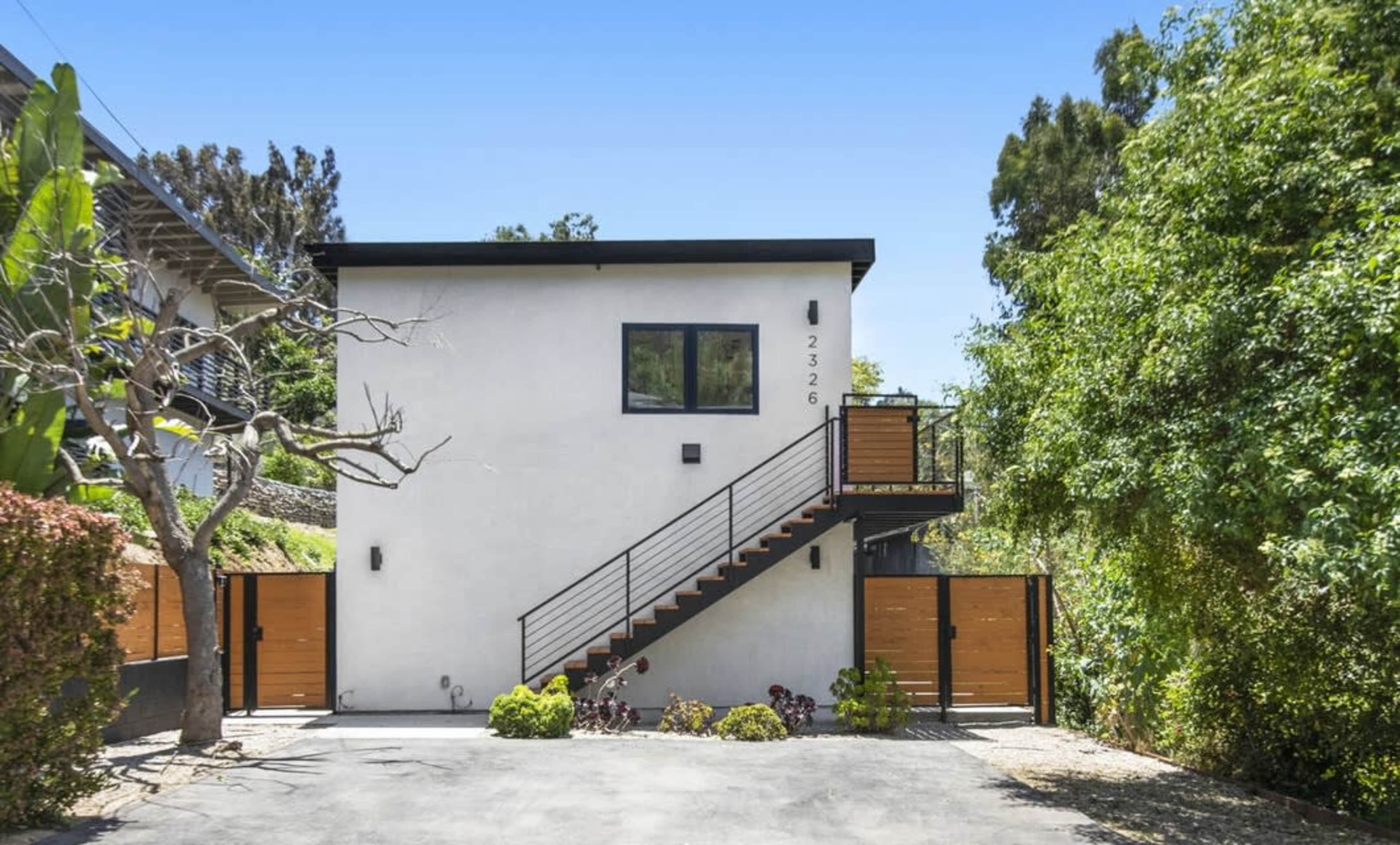 The image shows a modern two-story house with a staircase leading to the upper level, surrounded by lush greenery and two wooden garage doors.