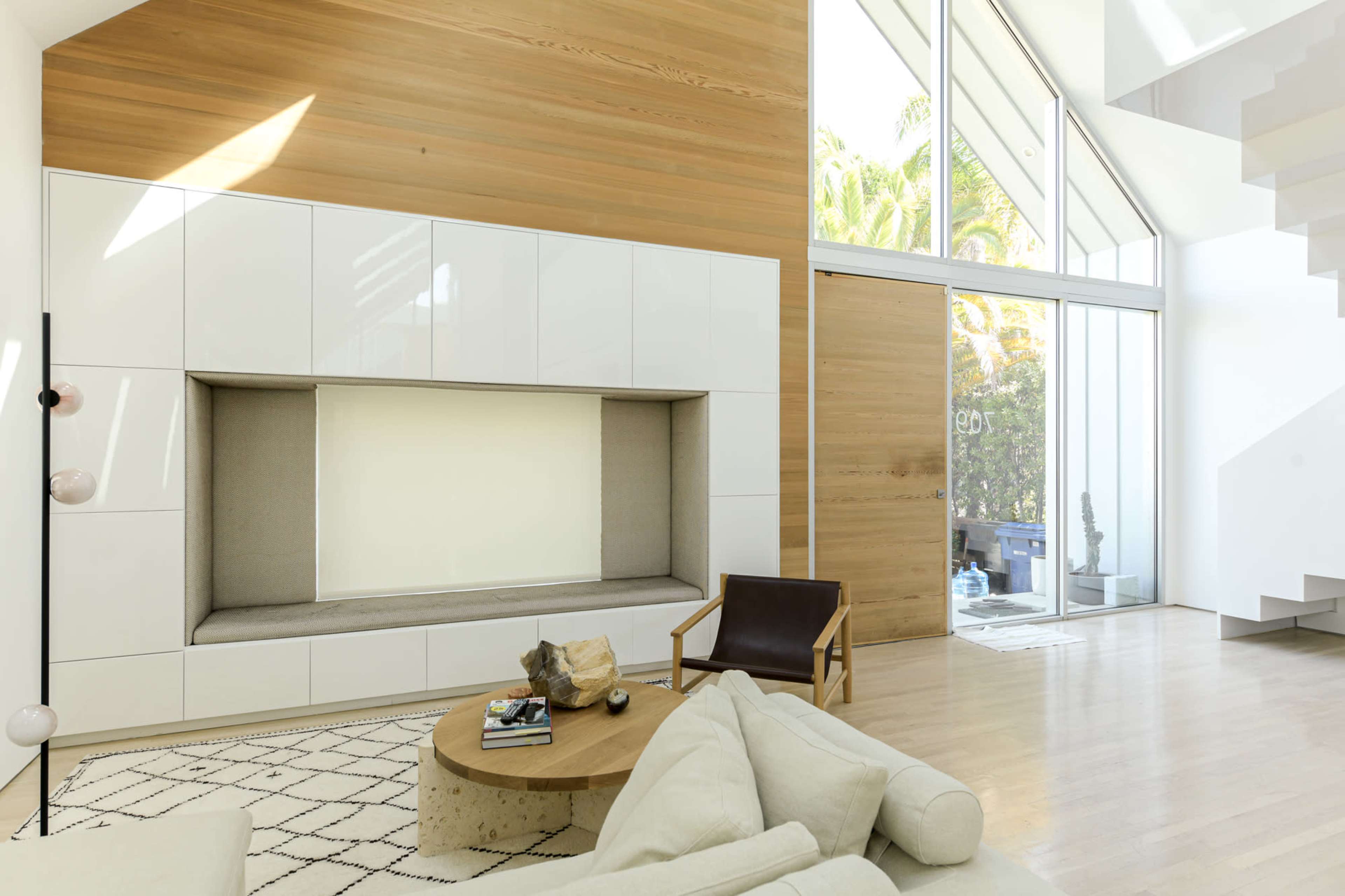 A bright living room features a large, minimalist entertainment unit, a circular coffee table, and a simple armchair, with large windows allowing sunlight to flood the space.