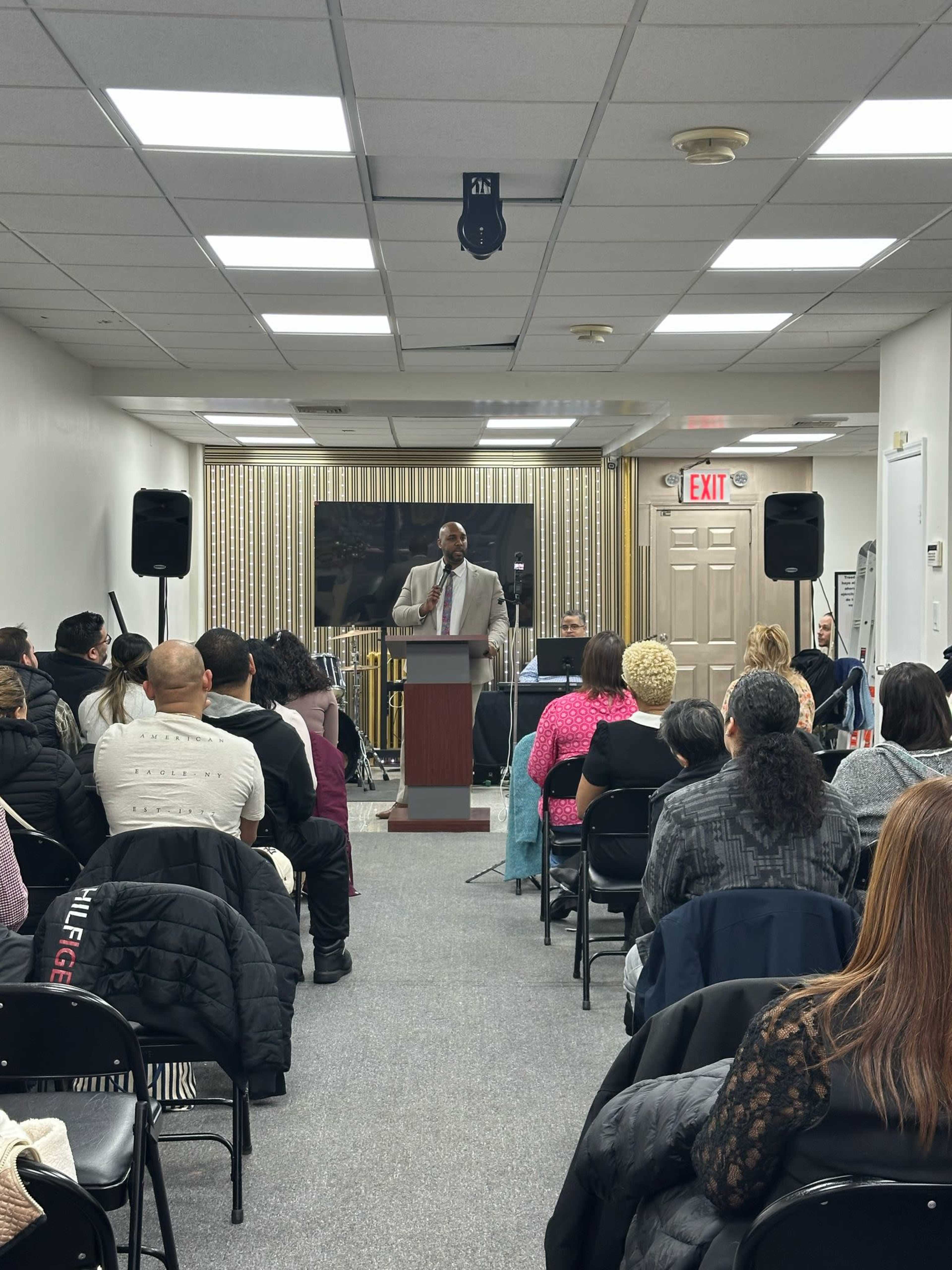 A speaker stands at a podium addressing an audience seated in a room with several chairs and a small stage setup.