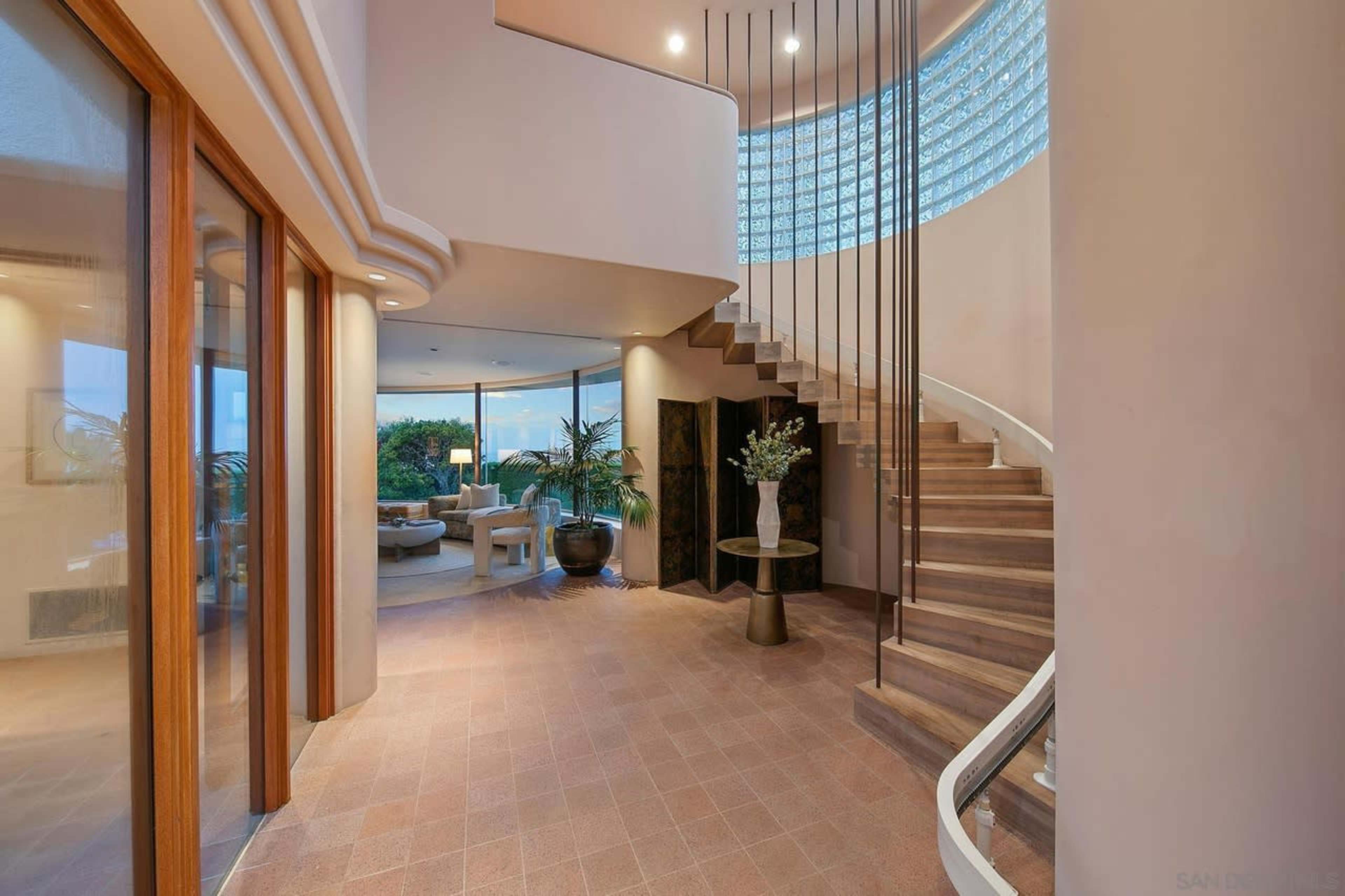 The image shows an elegant entryway with a spiral staircase, large glass block windows, and potted plants, leading into a spacious living area.