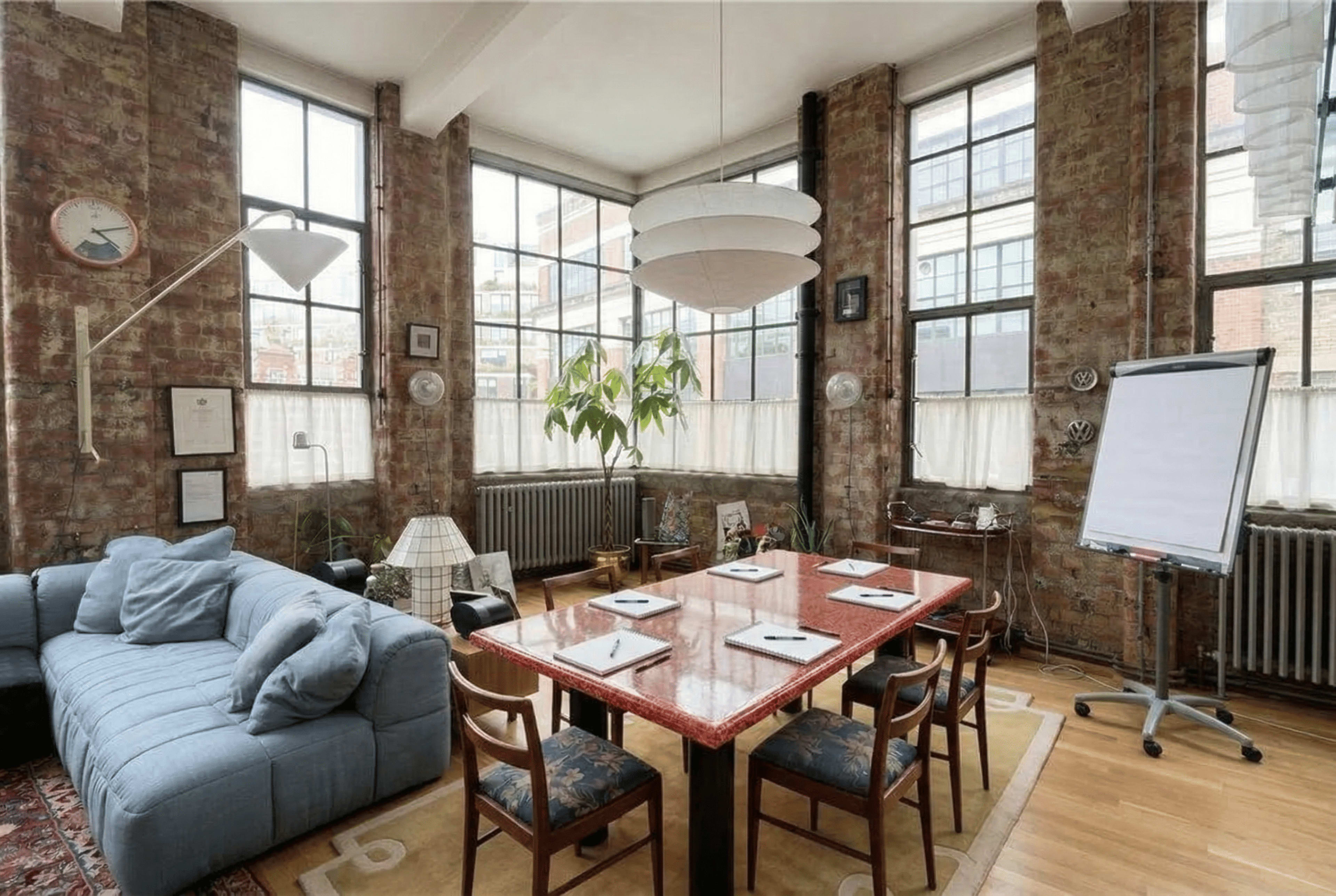 The image shows a spacious meeting room with a large red table surrounded by wooden chairs, illuminated by natural light from large windows, and featuring exposed brick walls and a potted plant.