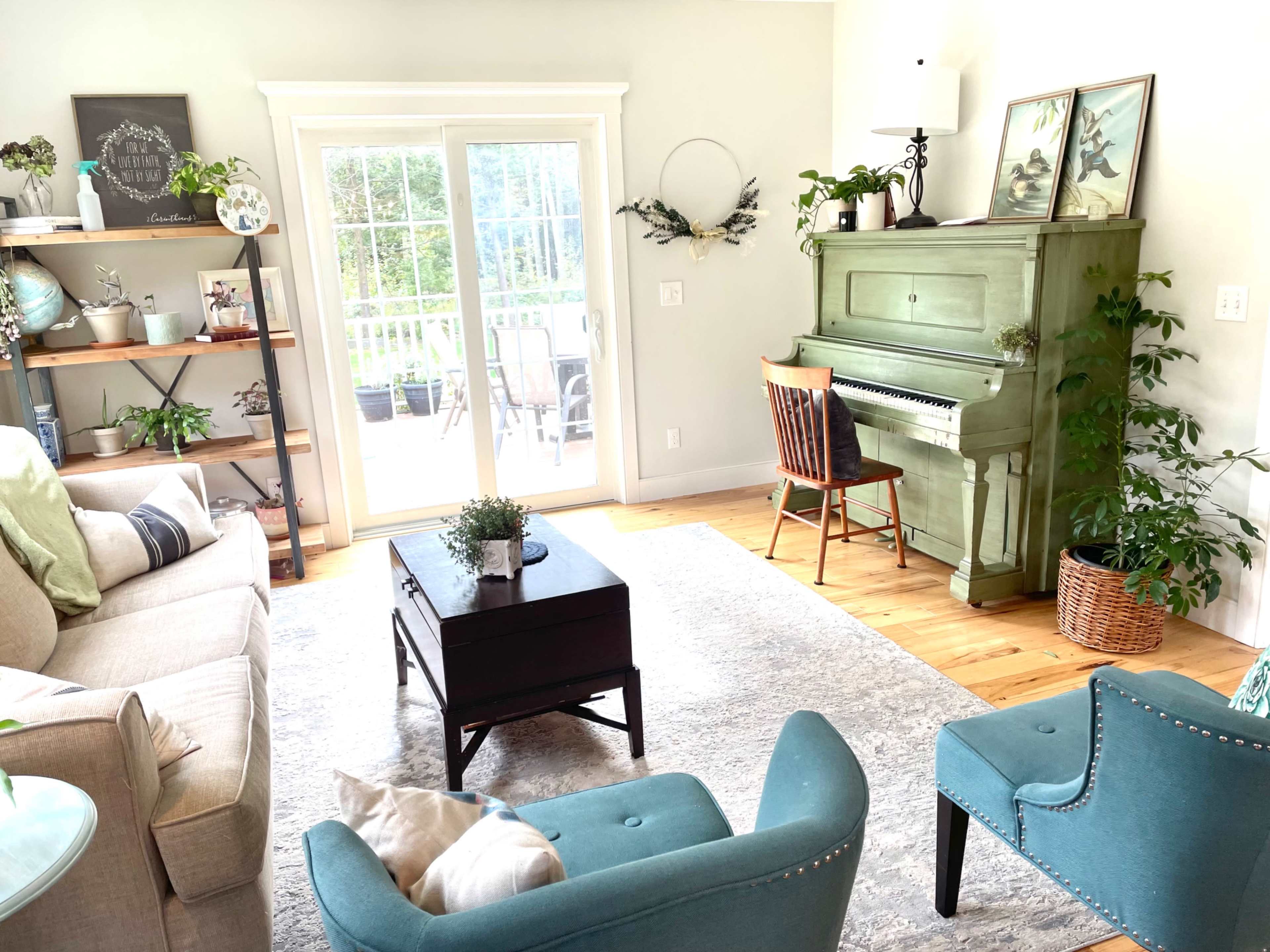 The living room features a green upright piano, a beige sofa, two teal chairs, and a wooden coffee table, with natural light coming through sliding glass doors.