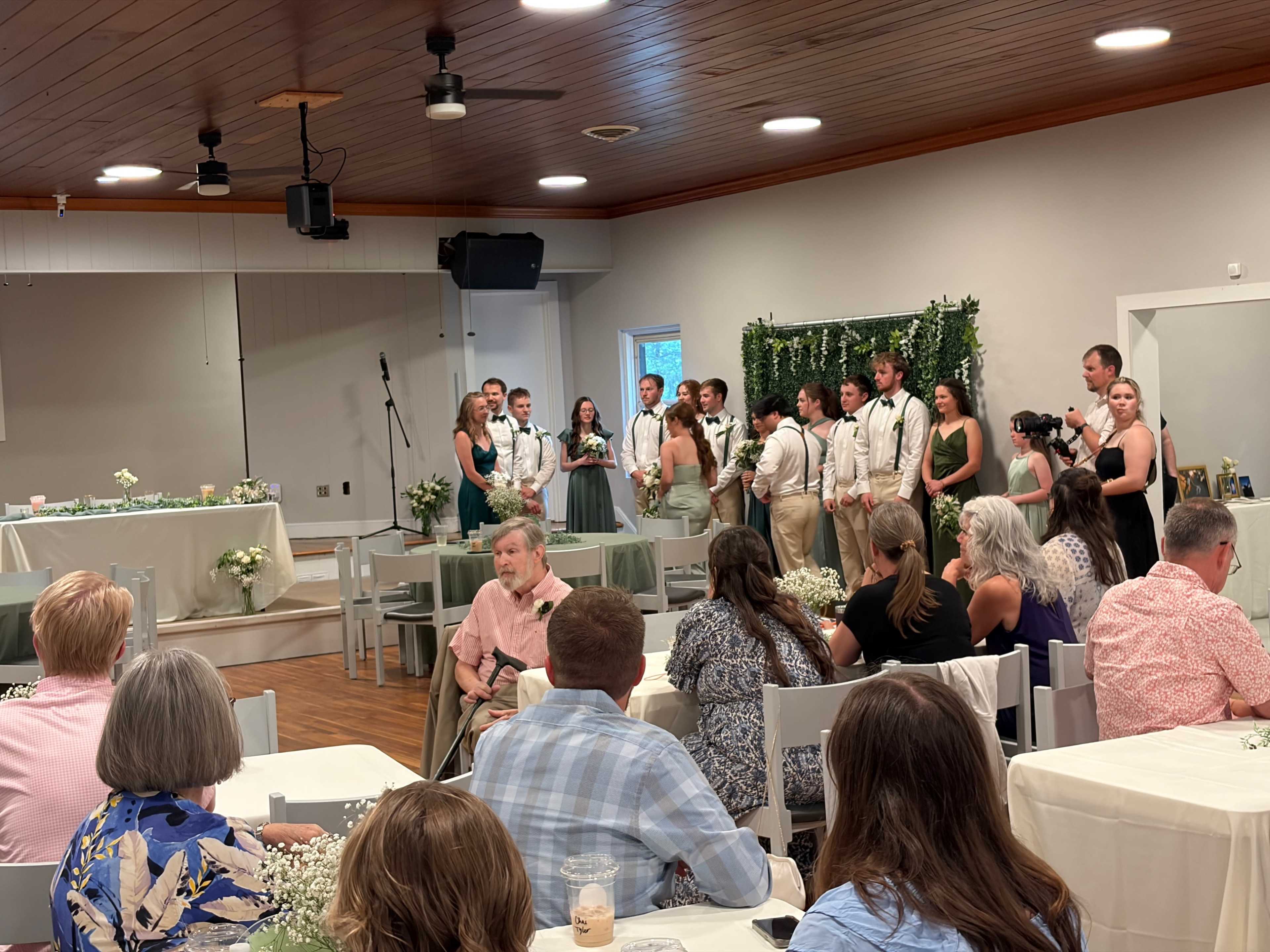 A wedding party stands in front of guests seated at tables in a decorated banquet hall.