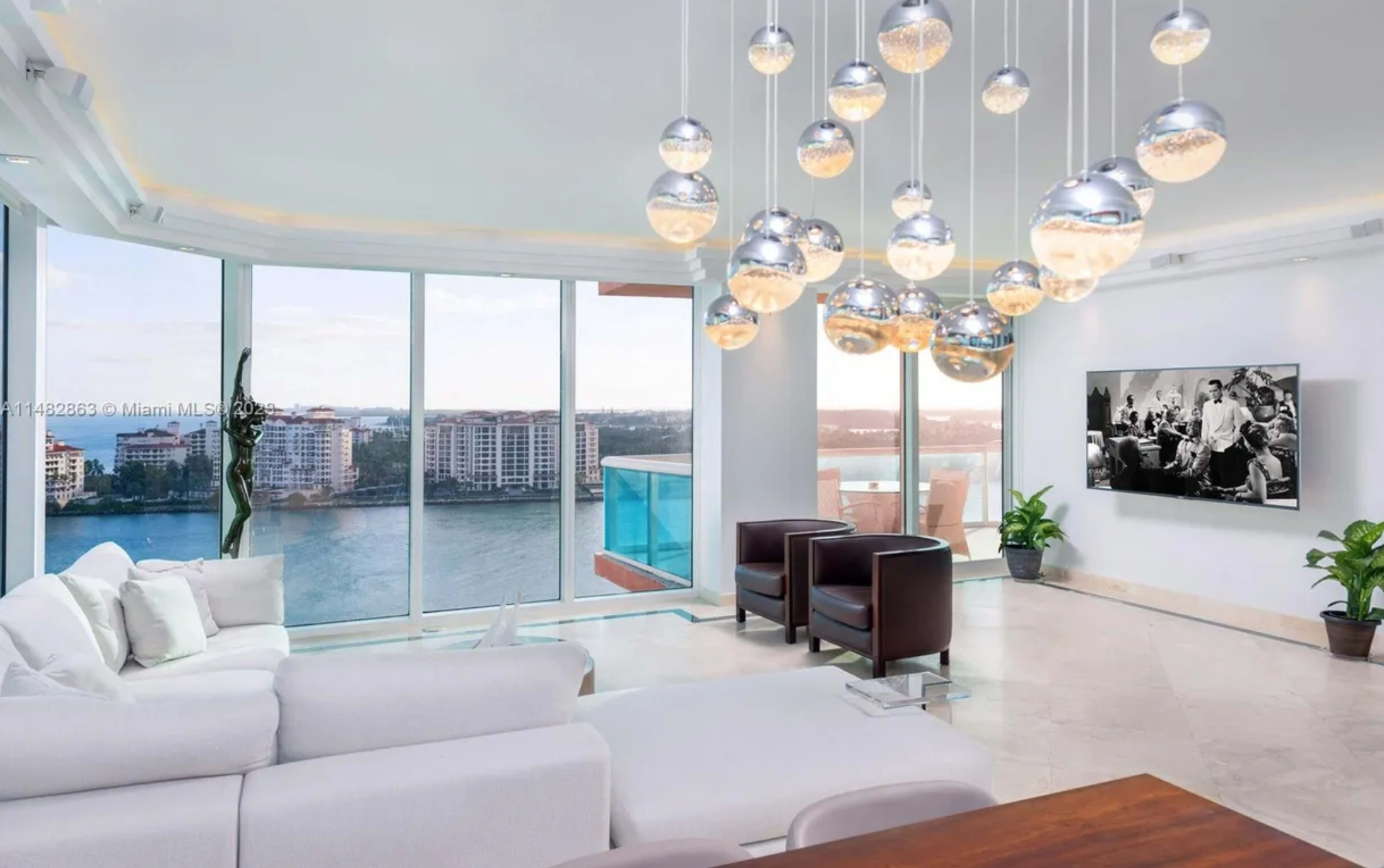 A modern living room with a large window displaying a waterfront view, two brown armchairs, a white sofa, a wooden dining table, and pendant lighting overhead.