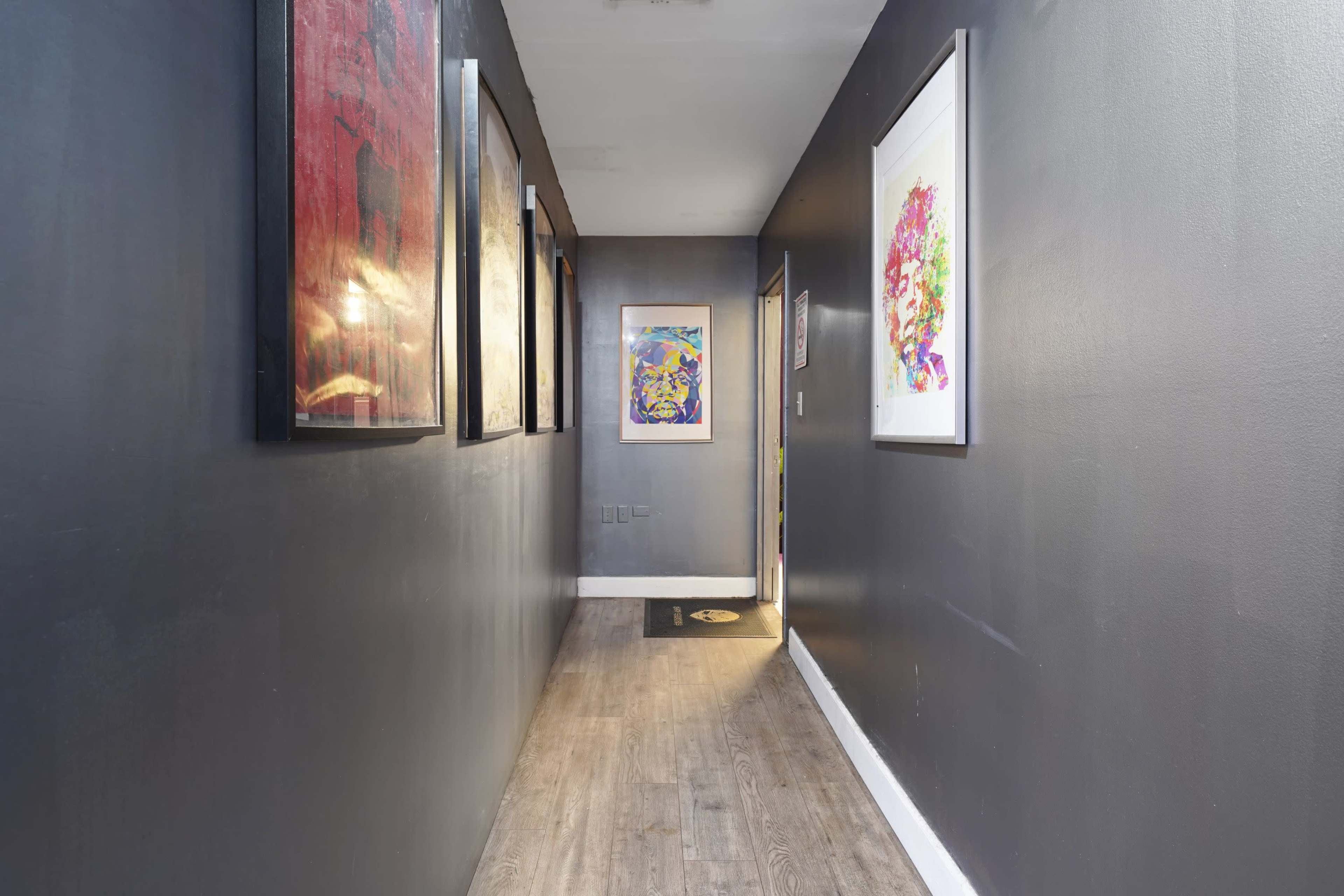 A narrow hallway features dark walls adorned with various framed artworks and wood flooring that leads to a door at the end.