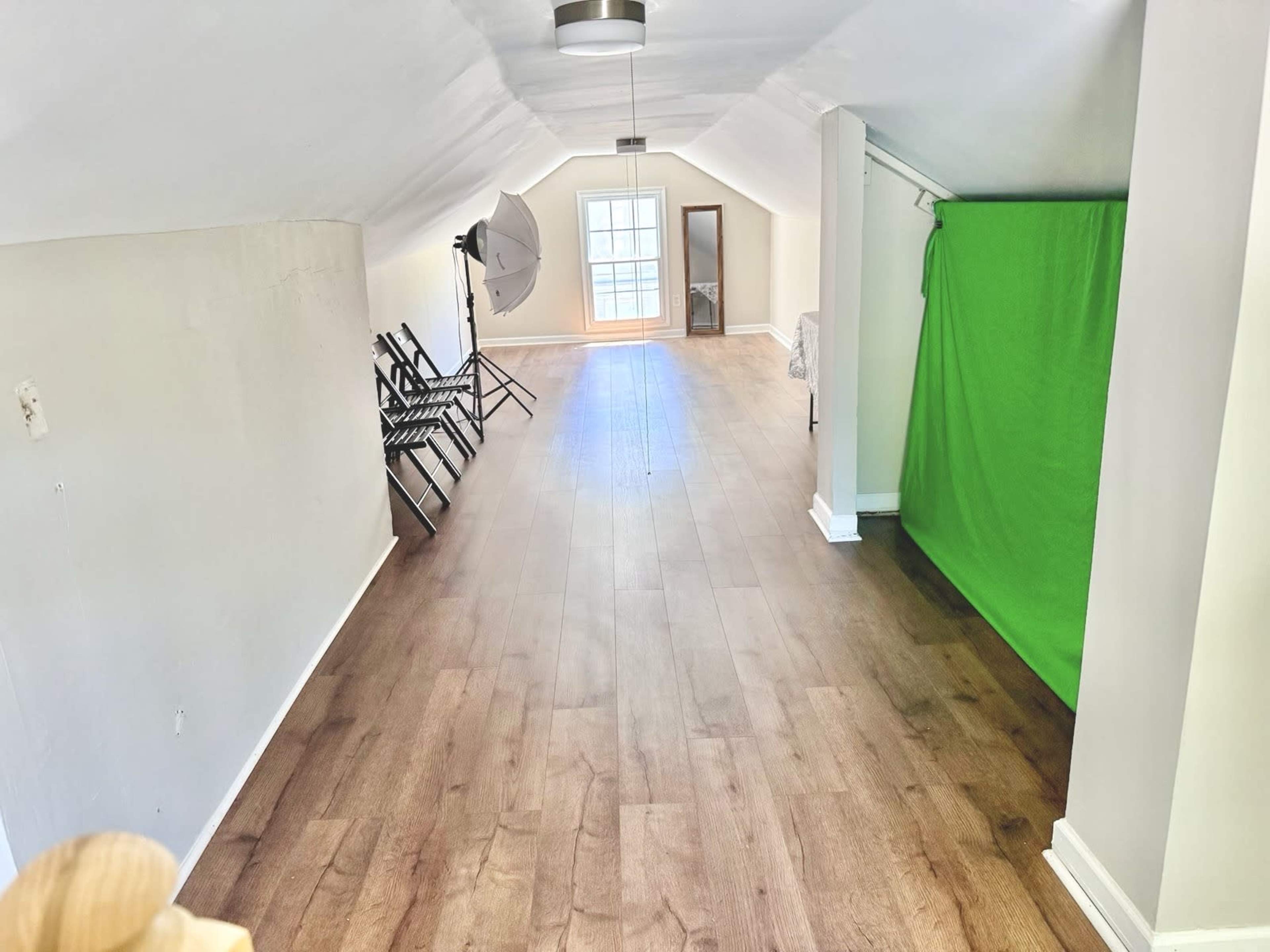 The image shows an attic-like space with wooden flooring, several folding chairs lined against one wall, a green backdrop in one corner, and a large window letting light in from the opposite side.