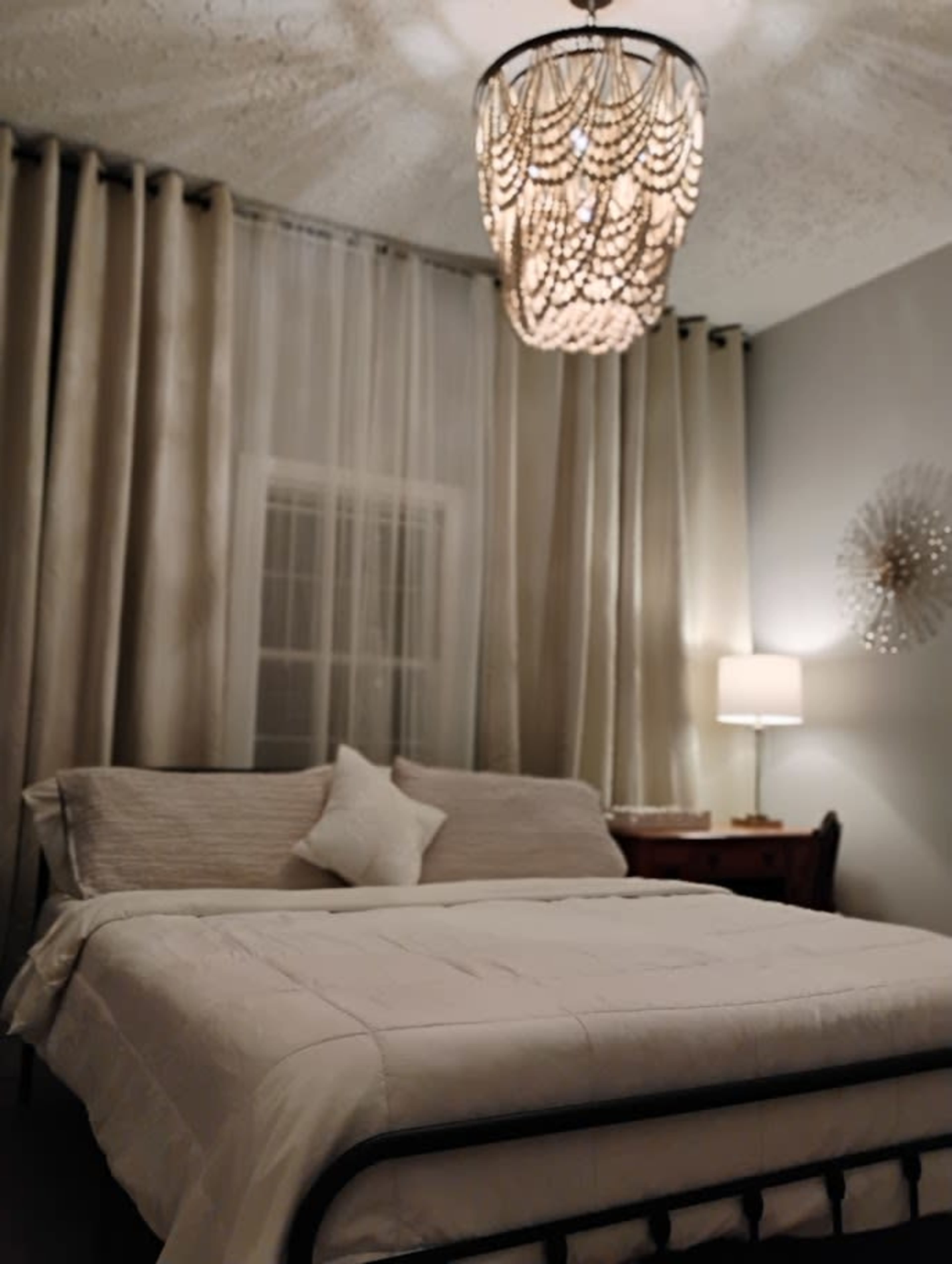 The image shows a bedroom featuring a bed with a white quilt, a decorative pillow, a bedside lamp, and a chandelier hanging from the ceiling.