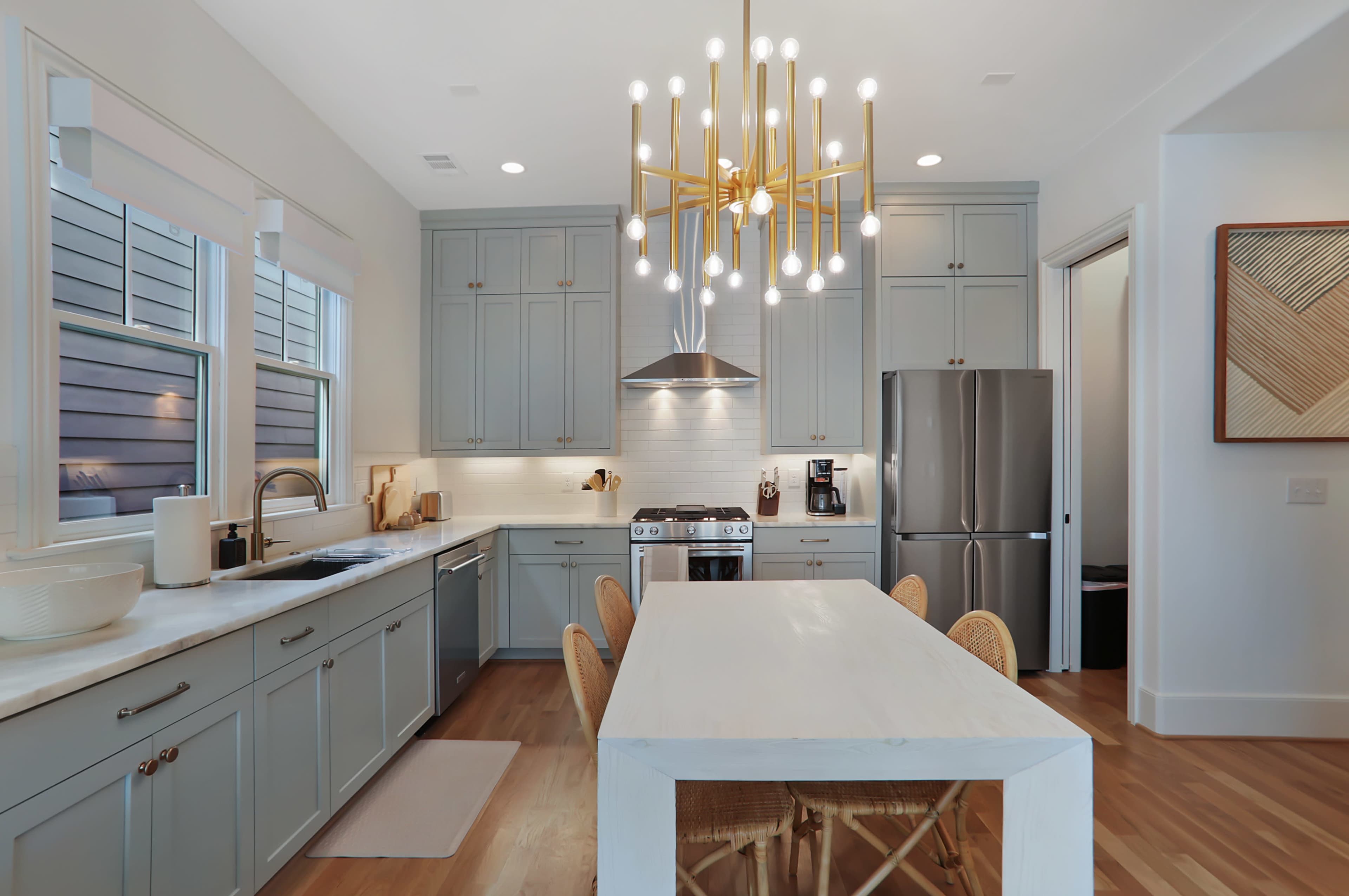A modern kitchen features blue cabinetry, a large white dining table, and a distinctive chandelier.