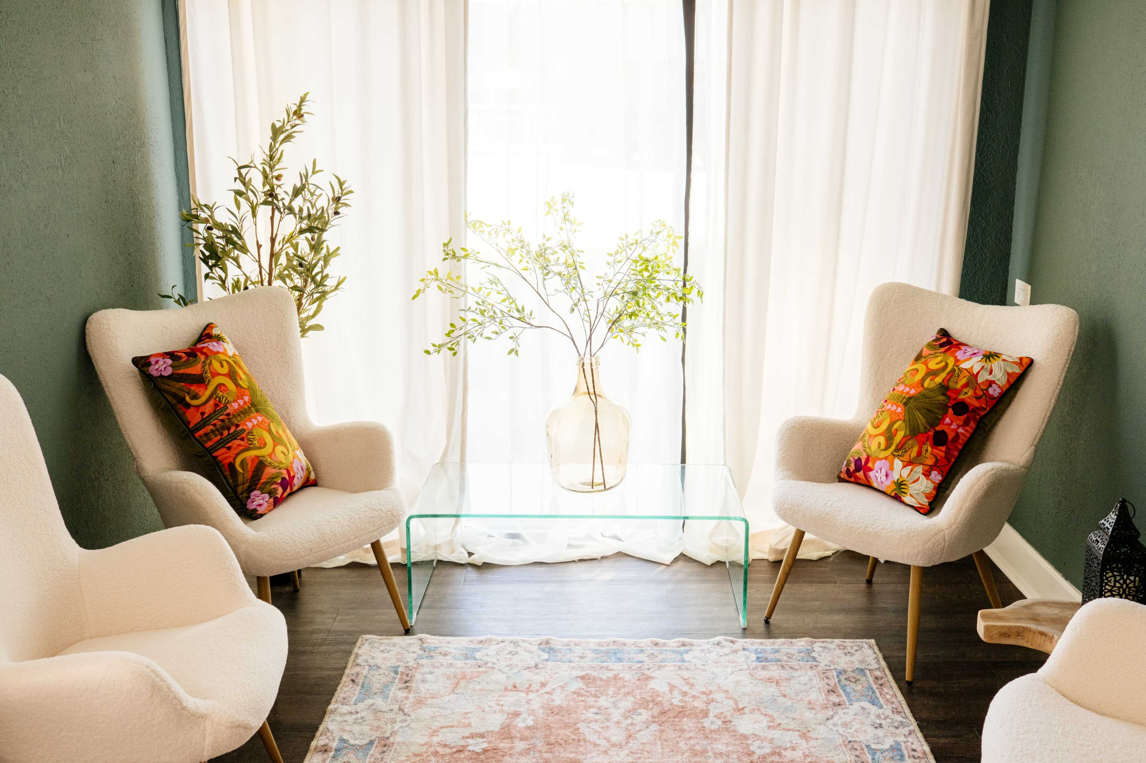 Two upholstered chairs with colorful pillows face a glass coffee table, positioned near a window with sheer white curtains, complemented by a vase of greenery.