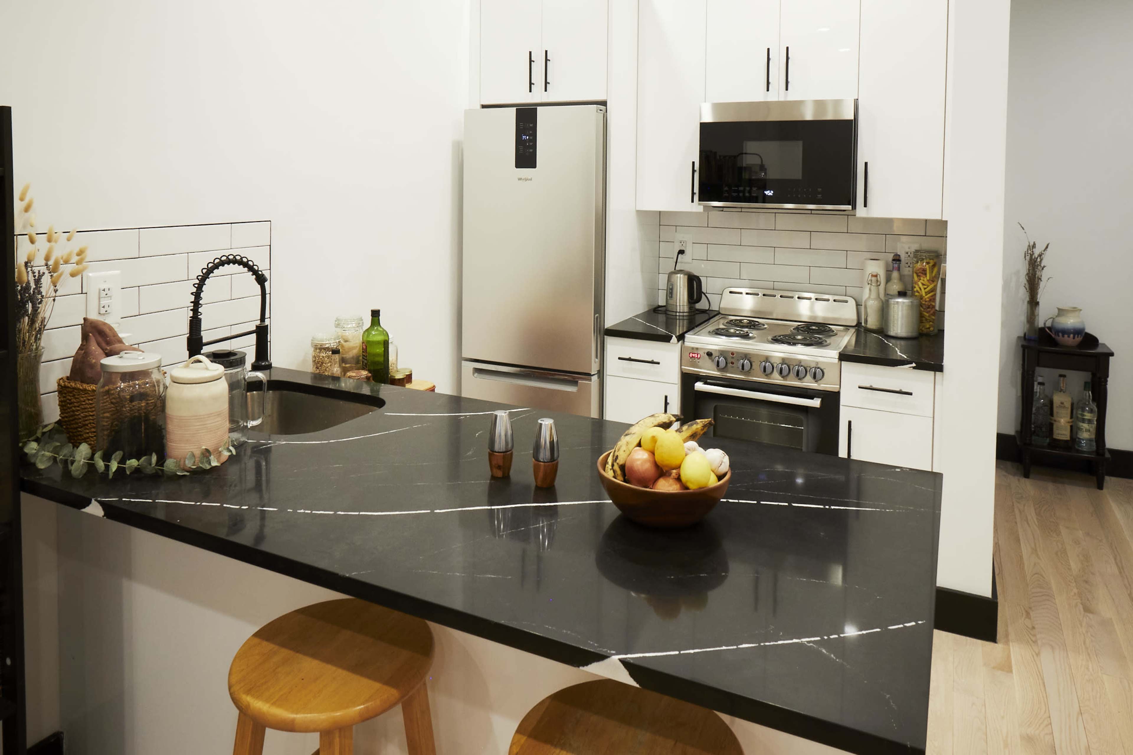 A modern kitchen features stainless steel appliances, dark countertops, a fruit bowl, and wooden stools at the island.