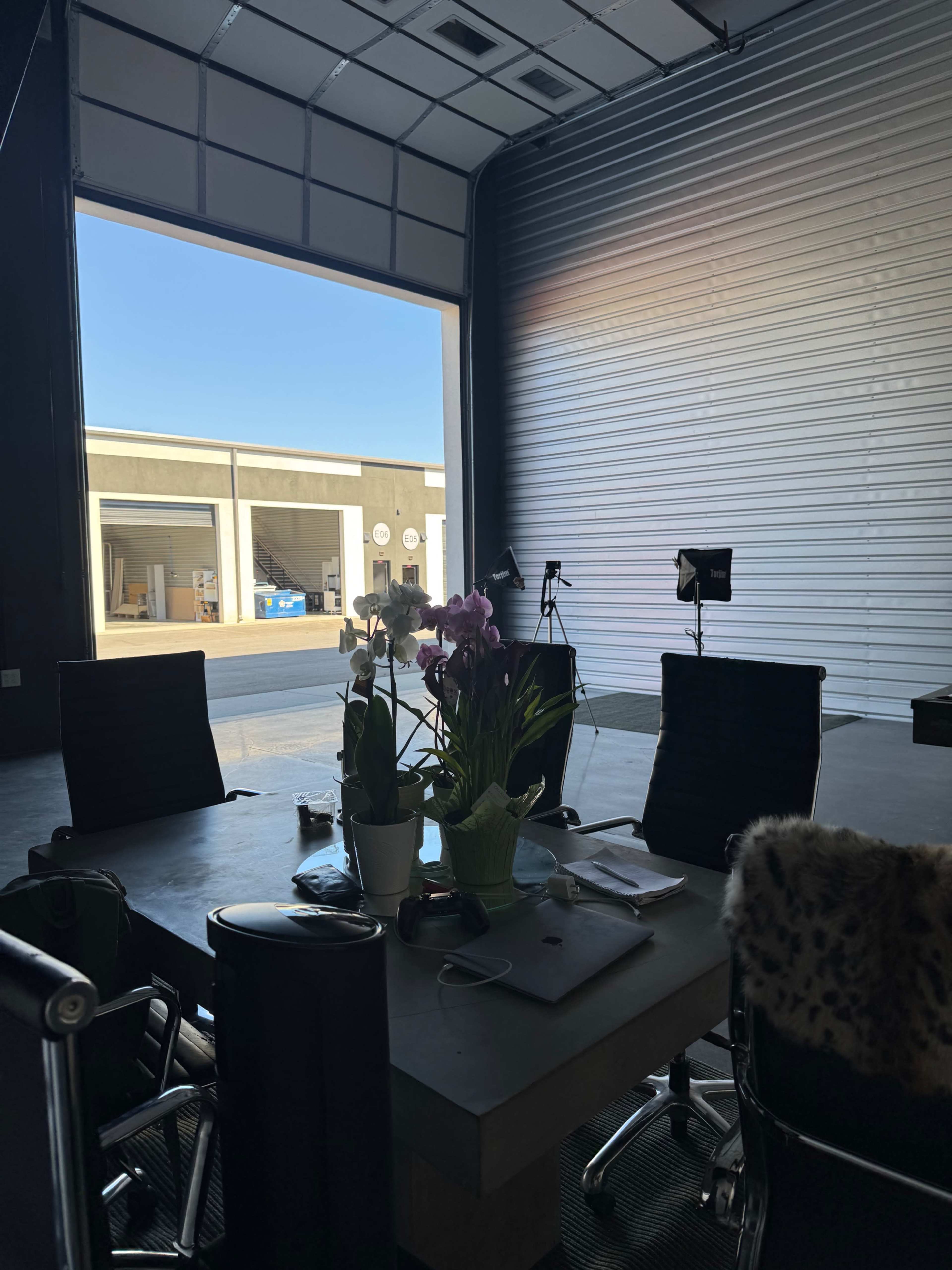 The image shows a spacious office area with a table and chairs, illuminated by natural light coming from an open garage door, revealing a view of an outdoor space with parked items.