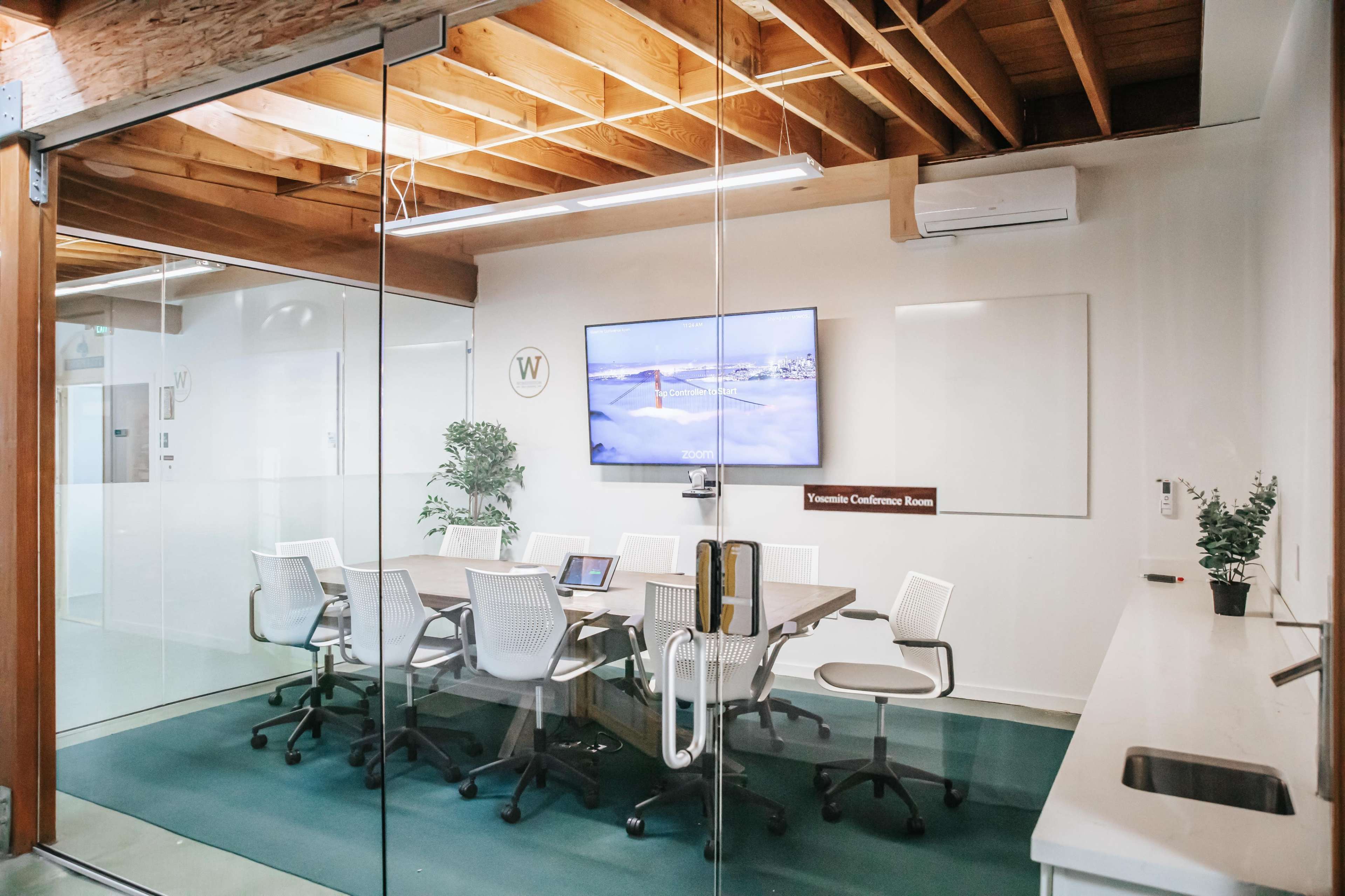 A modern conference room with a glass wall, a large screen on the wall, a table surrounded by six white chairs, and a small counter with a sink.