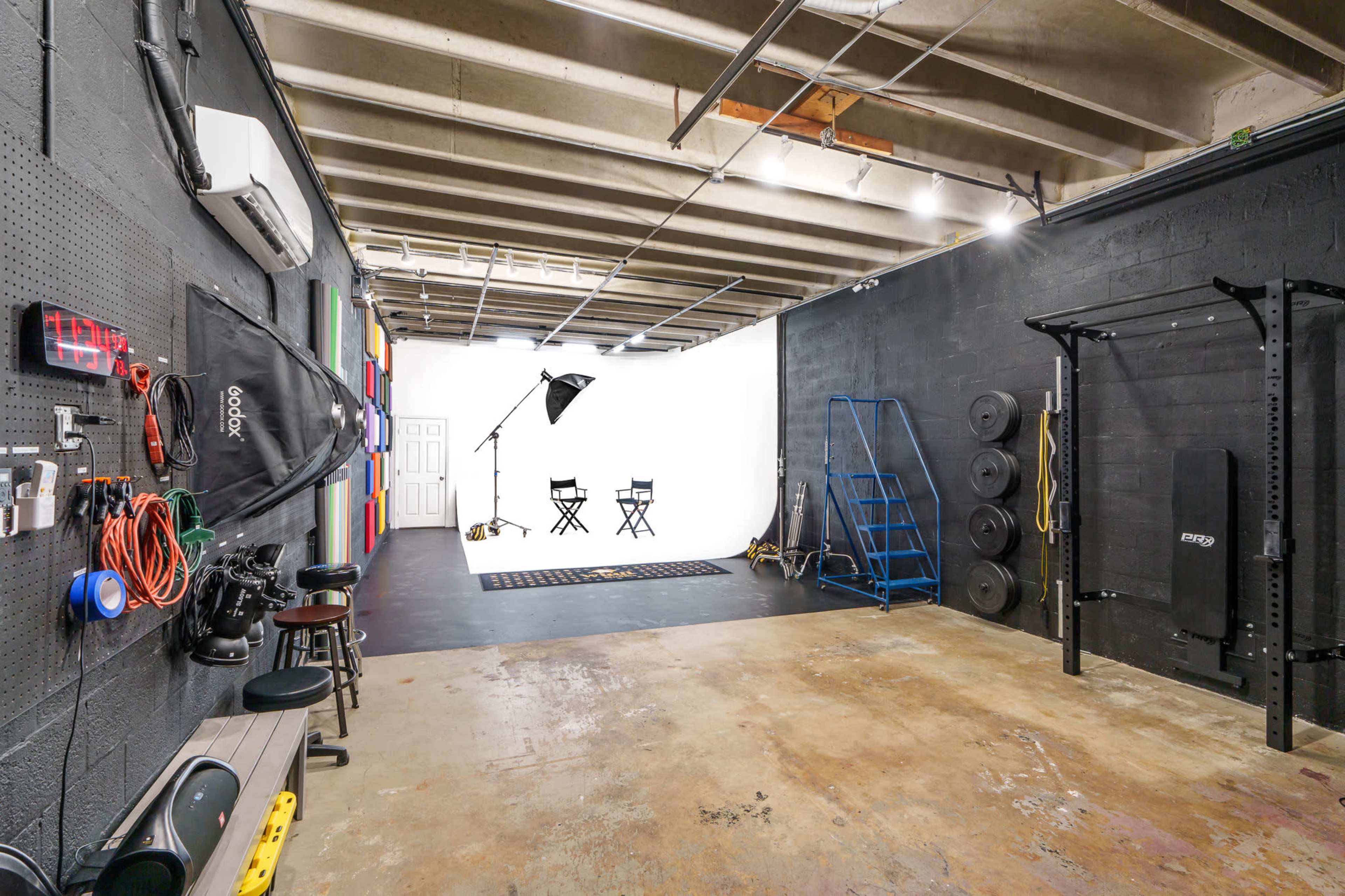 A spacious gym or studio area features black walls, exercise equipment, and a white backdrop with two directors' chairs.