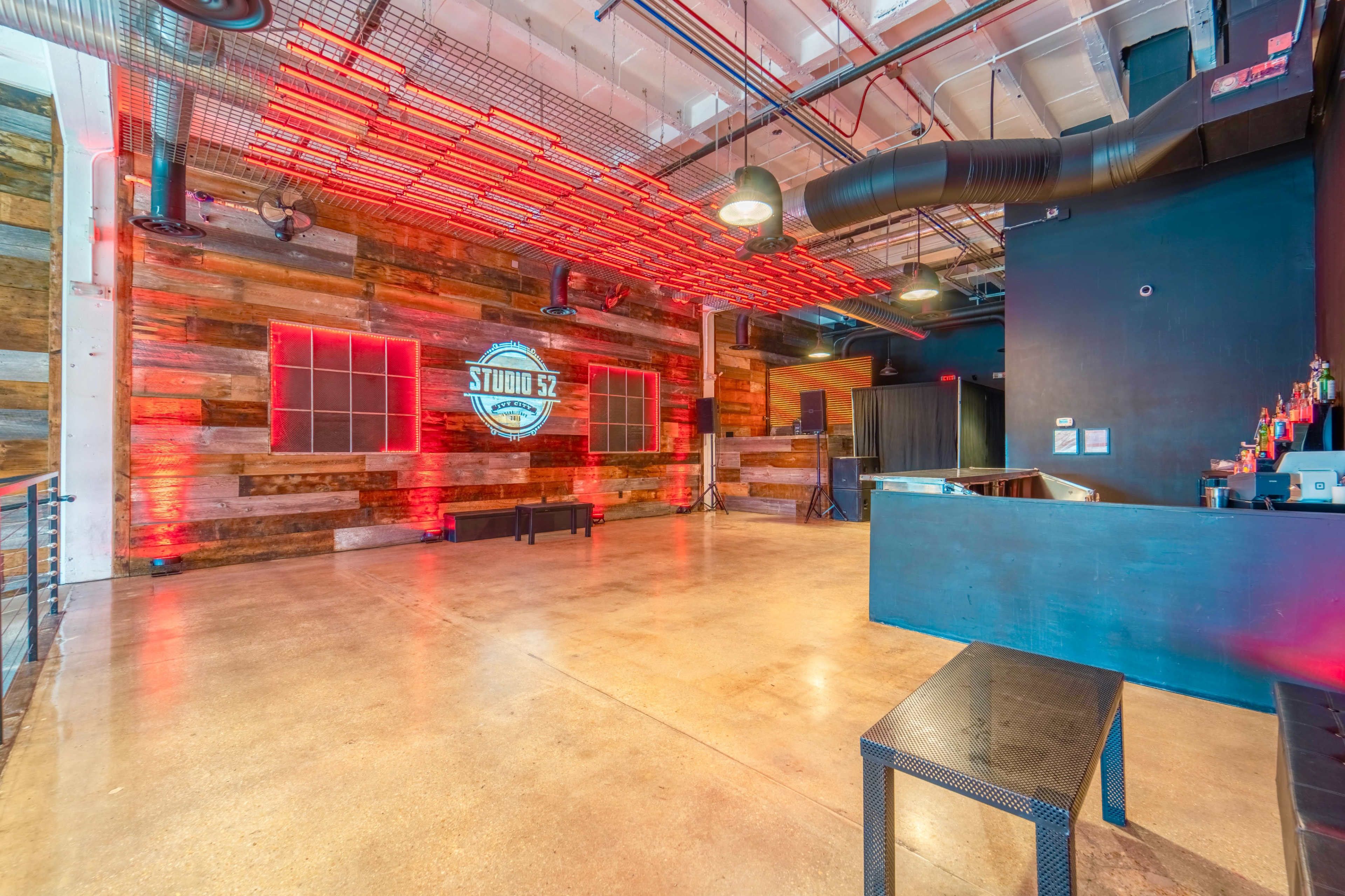 A spacious event venue with wooden-paneled walls, ambient red lighting, and a bar area against a black backdrop.