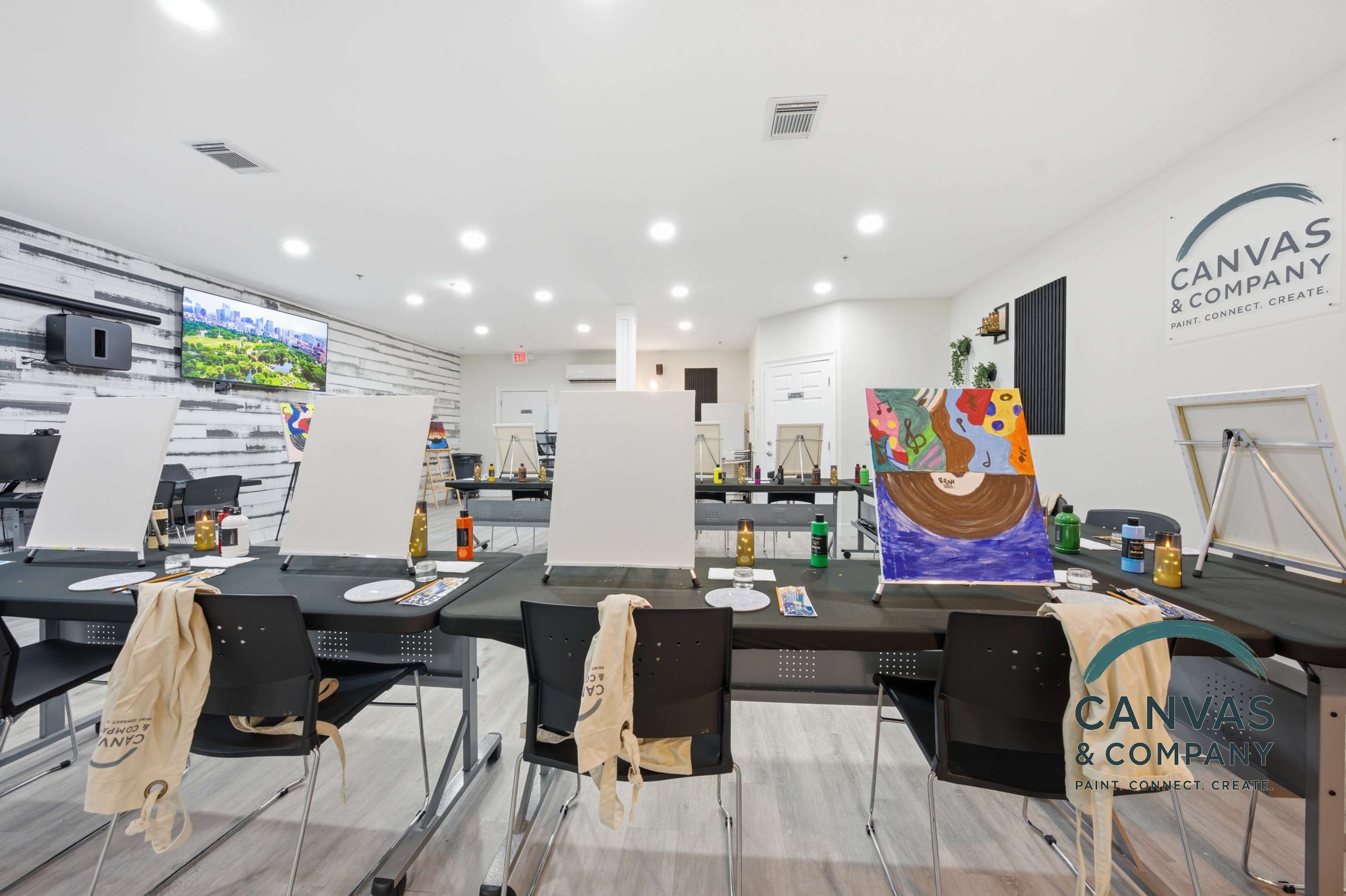 The image shows an art studio with several easels set up at black tables, each prepared with paint supplies and blank canvases, under bright lighting.
