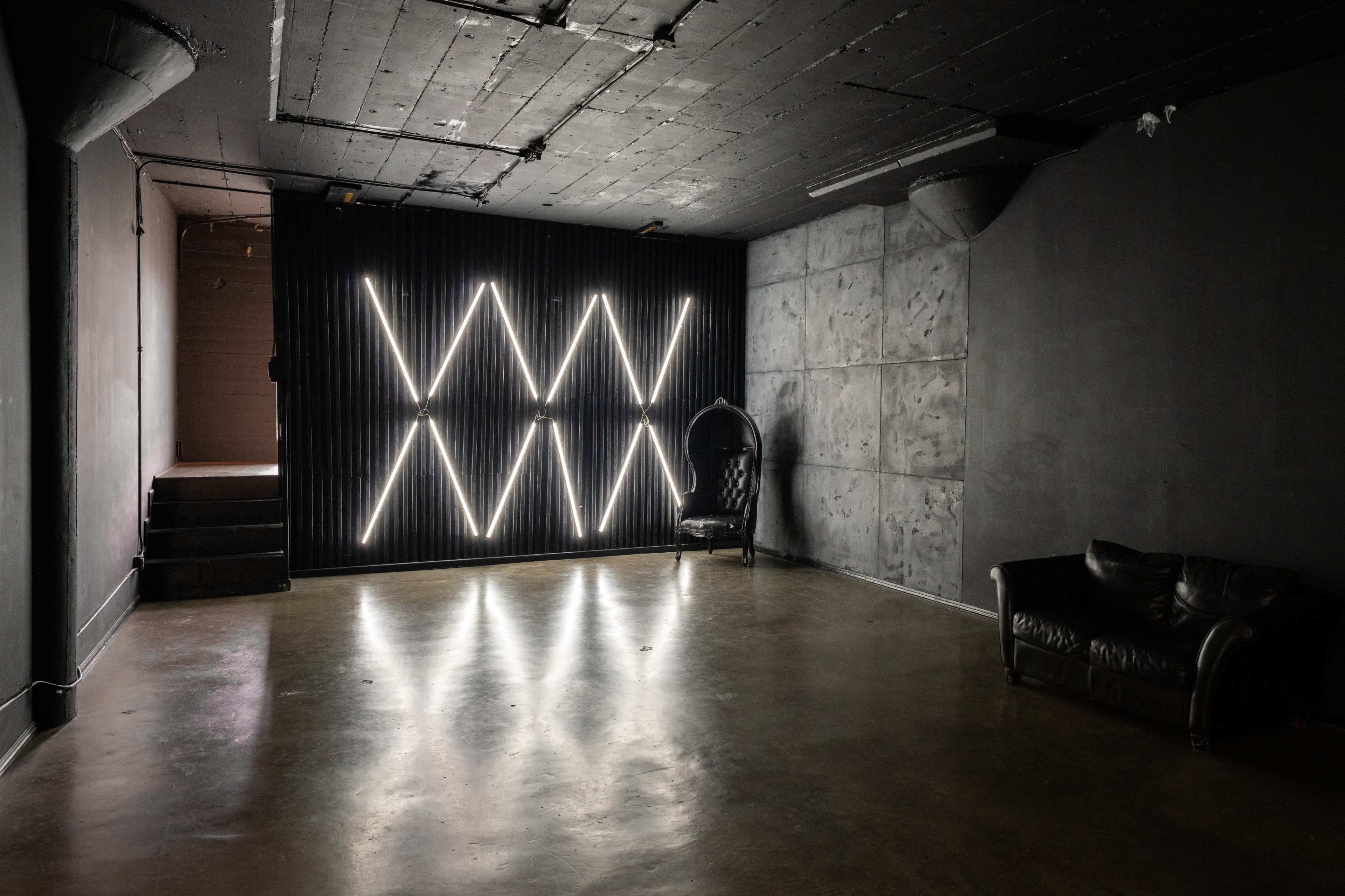 The image shows an empty industrial-style room with concrete walls, a dark floor, and minimalist lighting fixtures arranged in a geometric pattern.