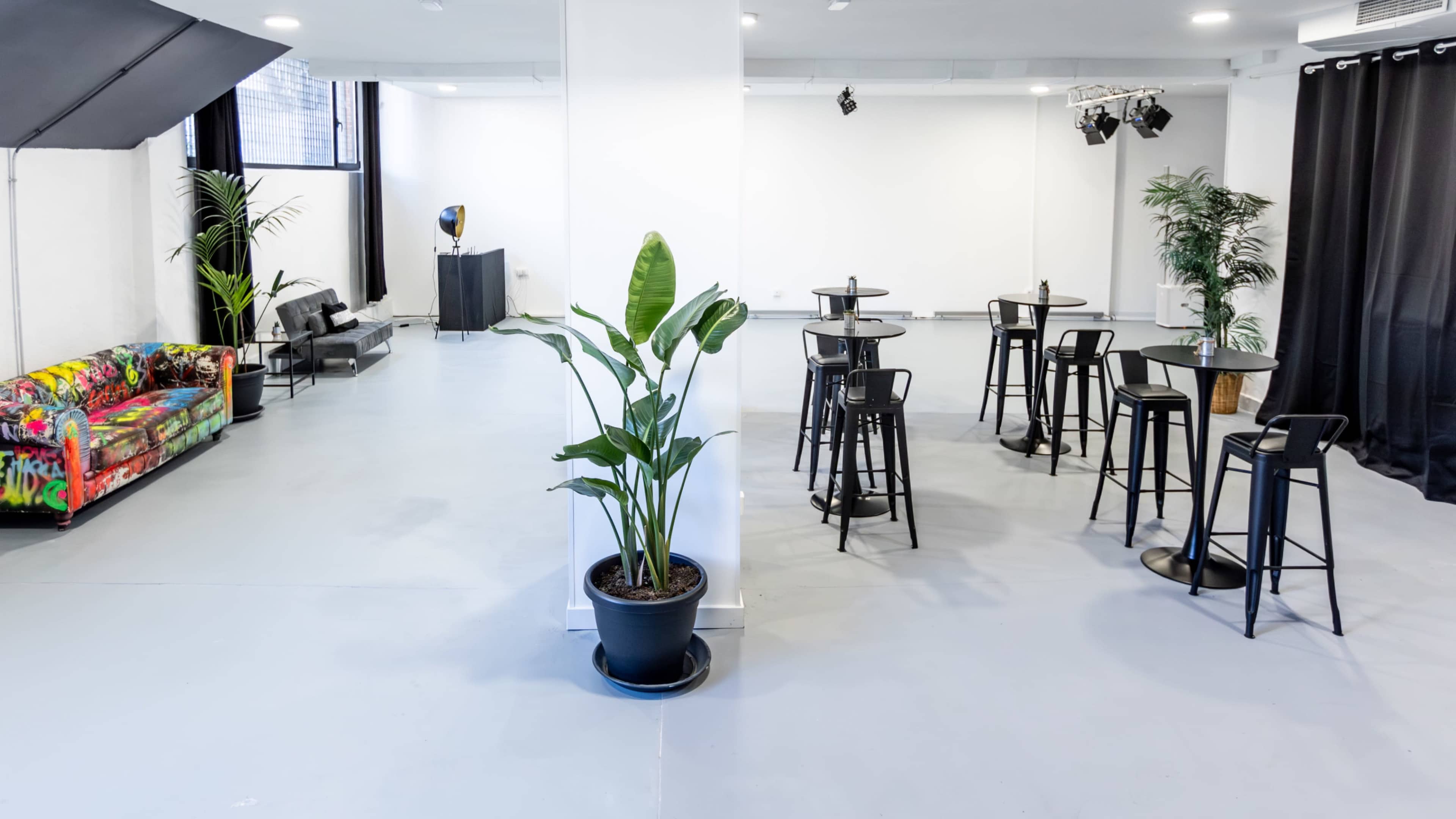 The image shows an open event space with colorful seating, tall tables, and potted plants, set in a well-lit room with white walls and a sound system in the background.