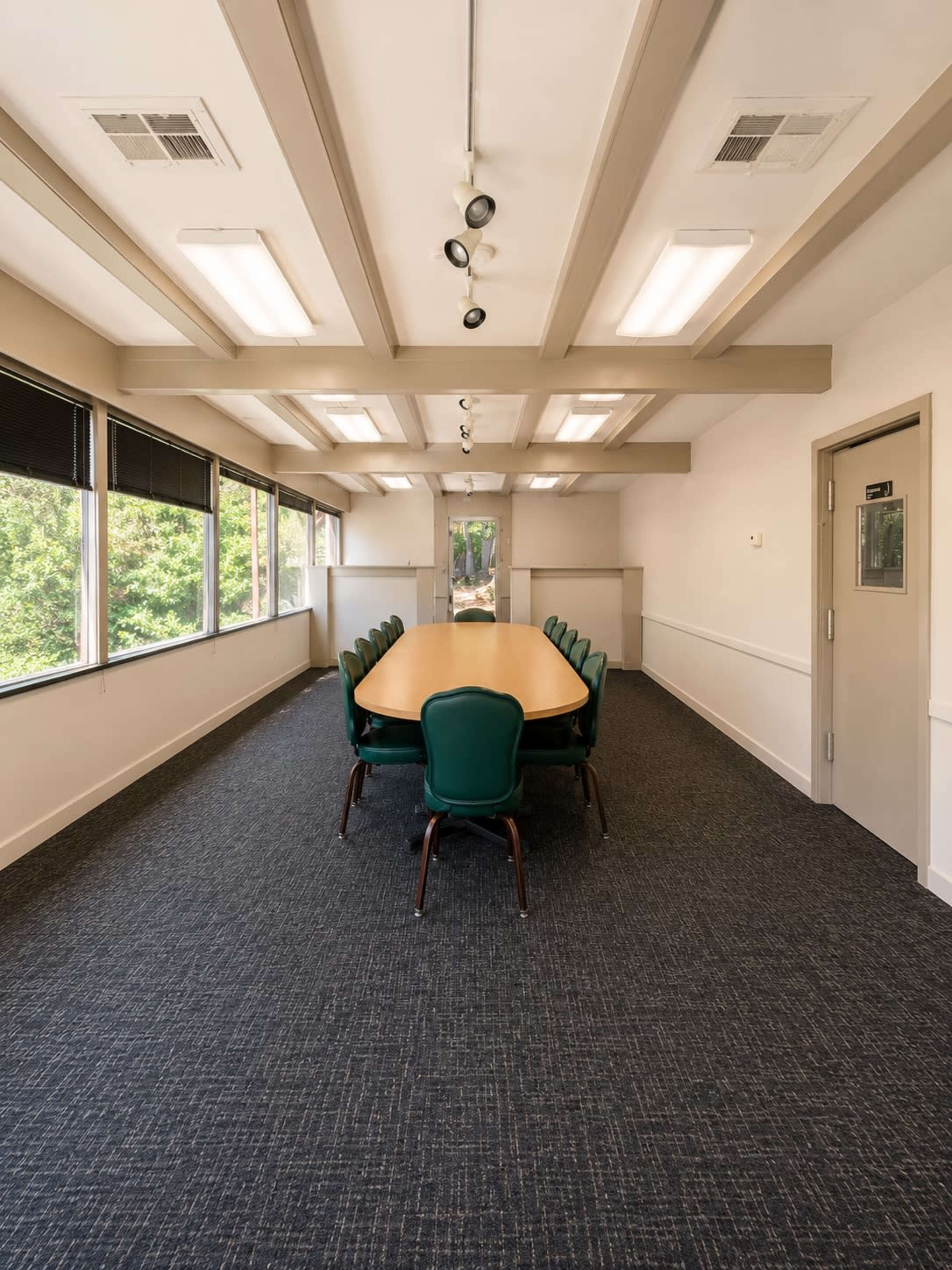 A conference room features a long wooden table surrounded by green chairs, with large windows letting in natural light.