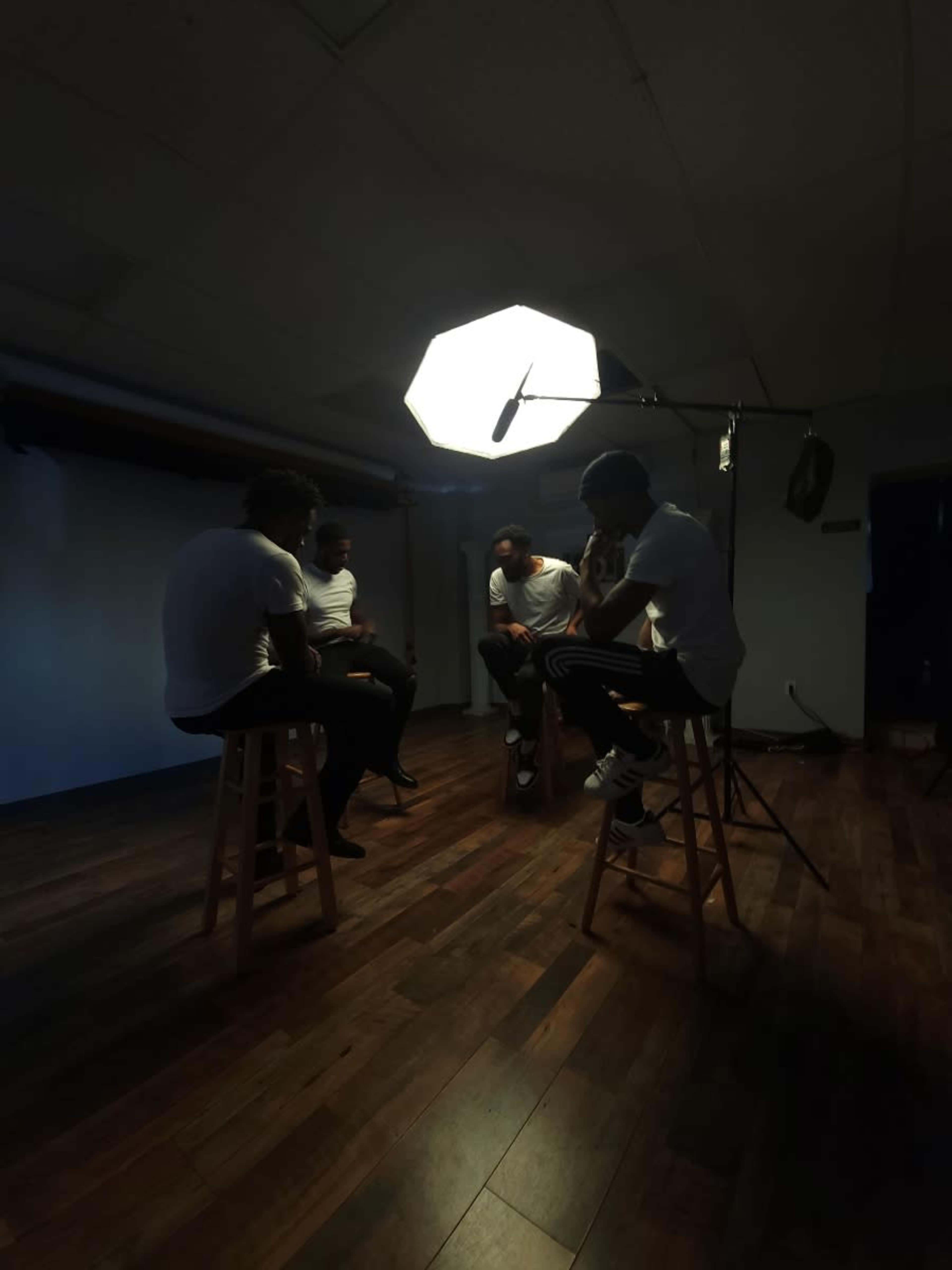 Four individuals dressed in white shirts are seated on stools in a dimly lit room, illuminated by a large softbox light.