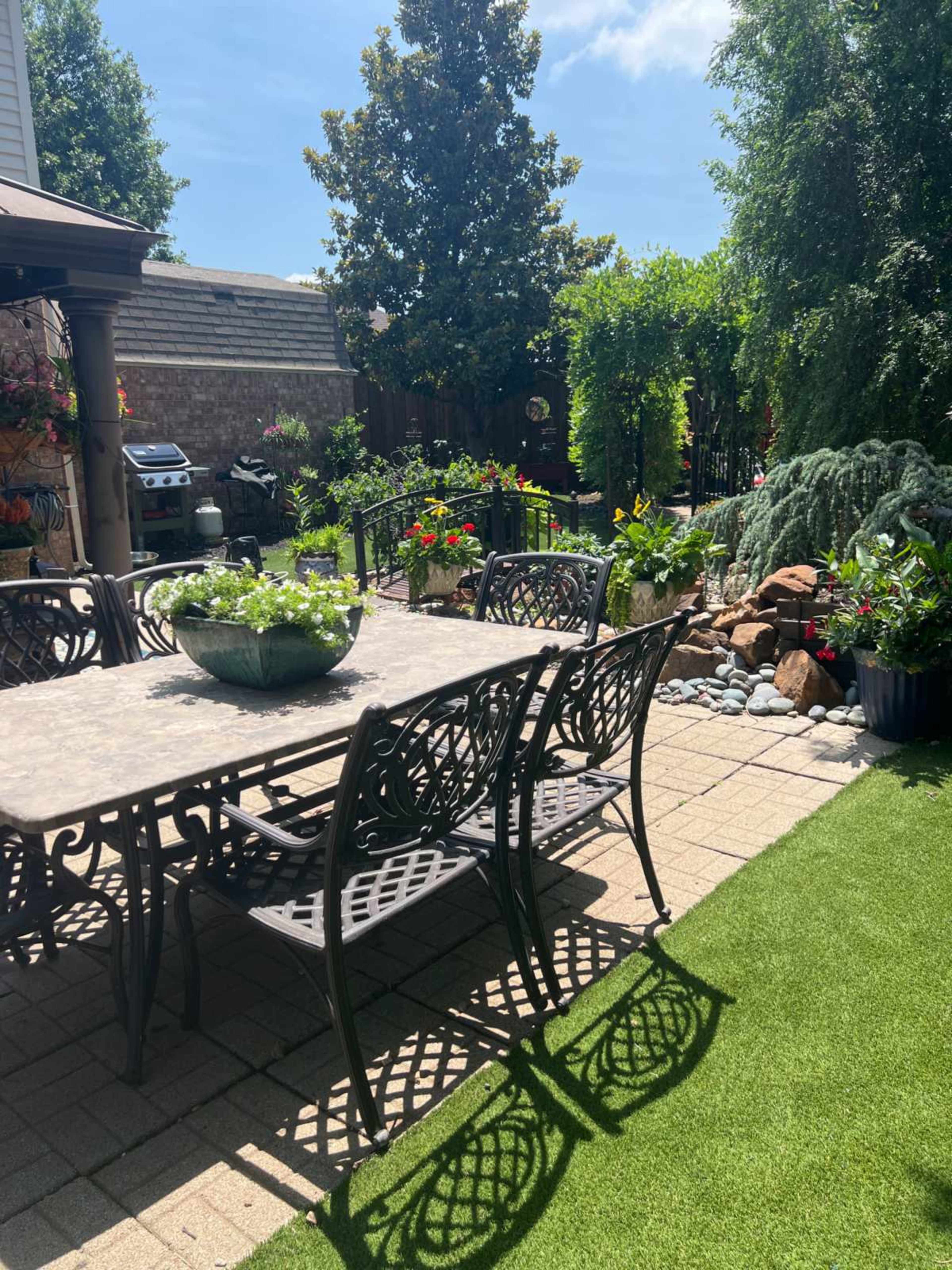 A patio area features a wrought iron dining table surrounded by chairs, with flowers and plants in various containers and a small rock garden in the background.