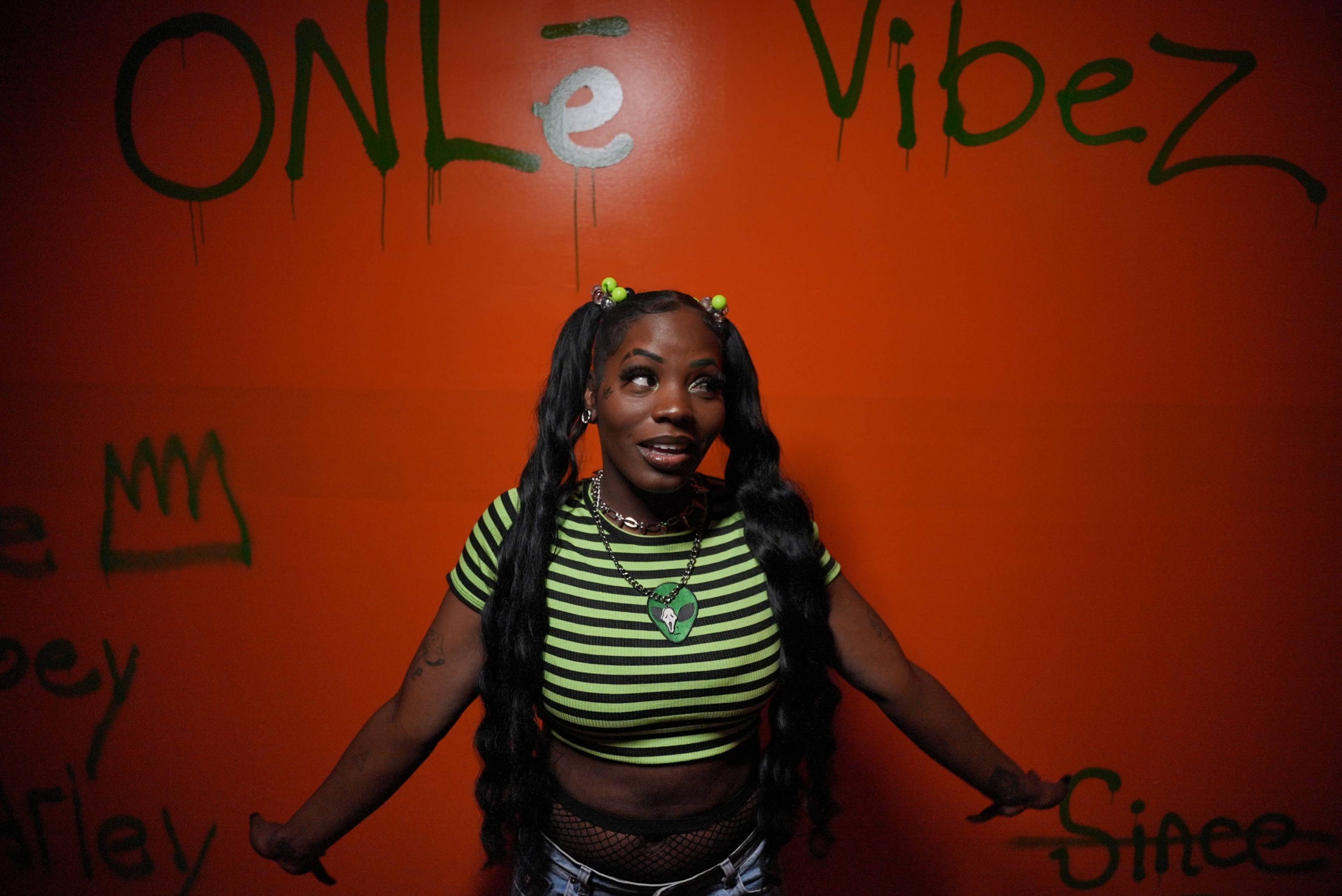 A woman with long braids stands against an orange wall with green graffiti, wearing a striped top and making a joyful gesture.