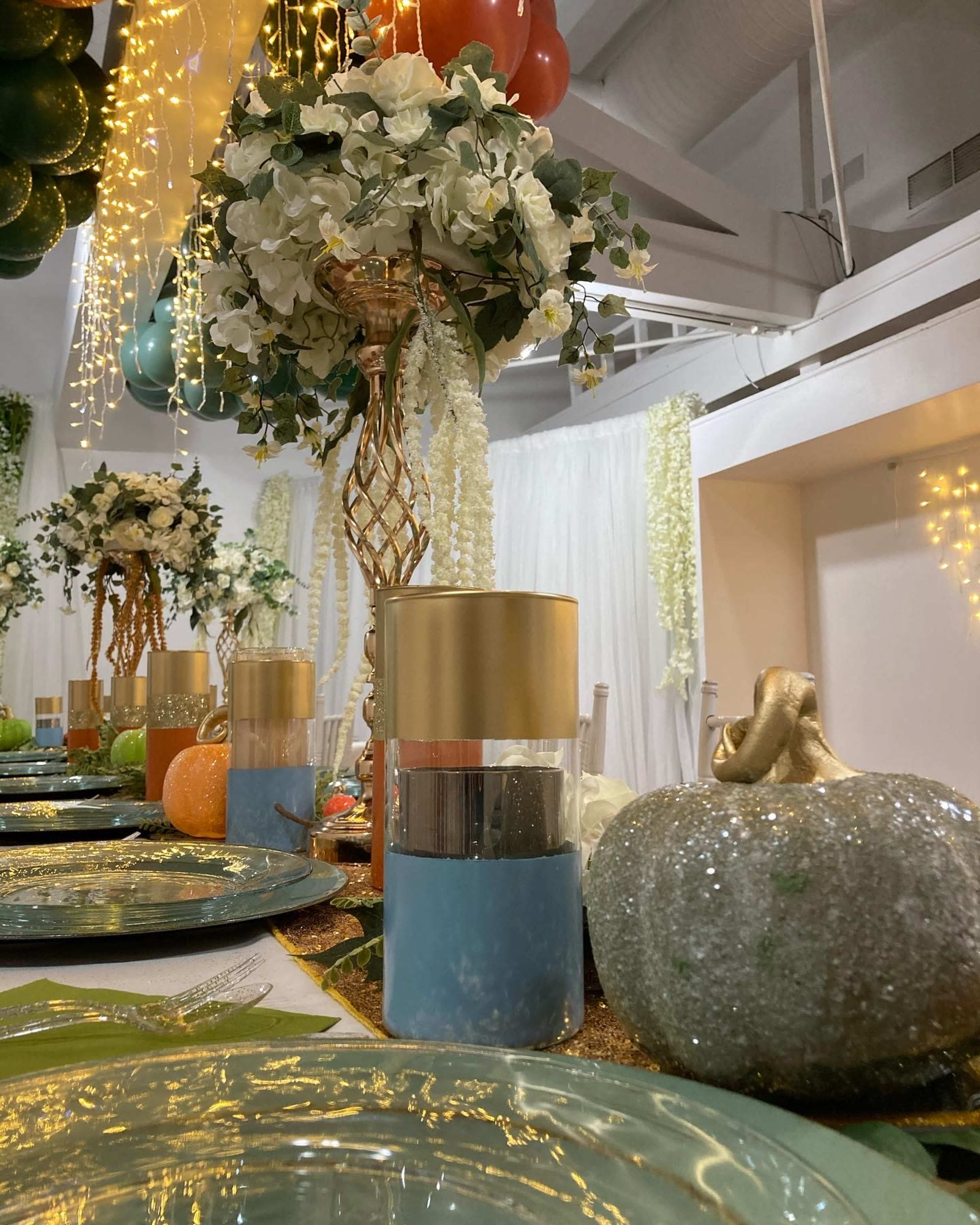 The image displays a elegantly arranged dining table with decorative centerpieces, including vases with flowers and sparkly pumpkins, under a backdrop of fairy lights.