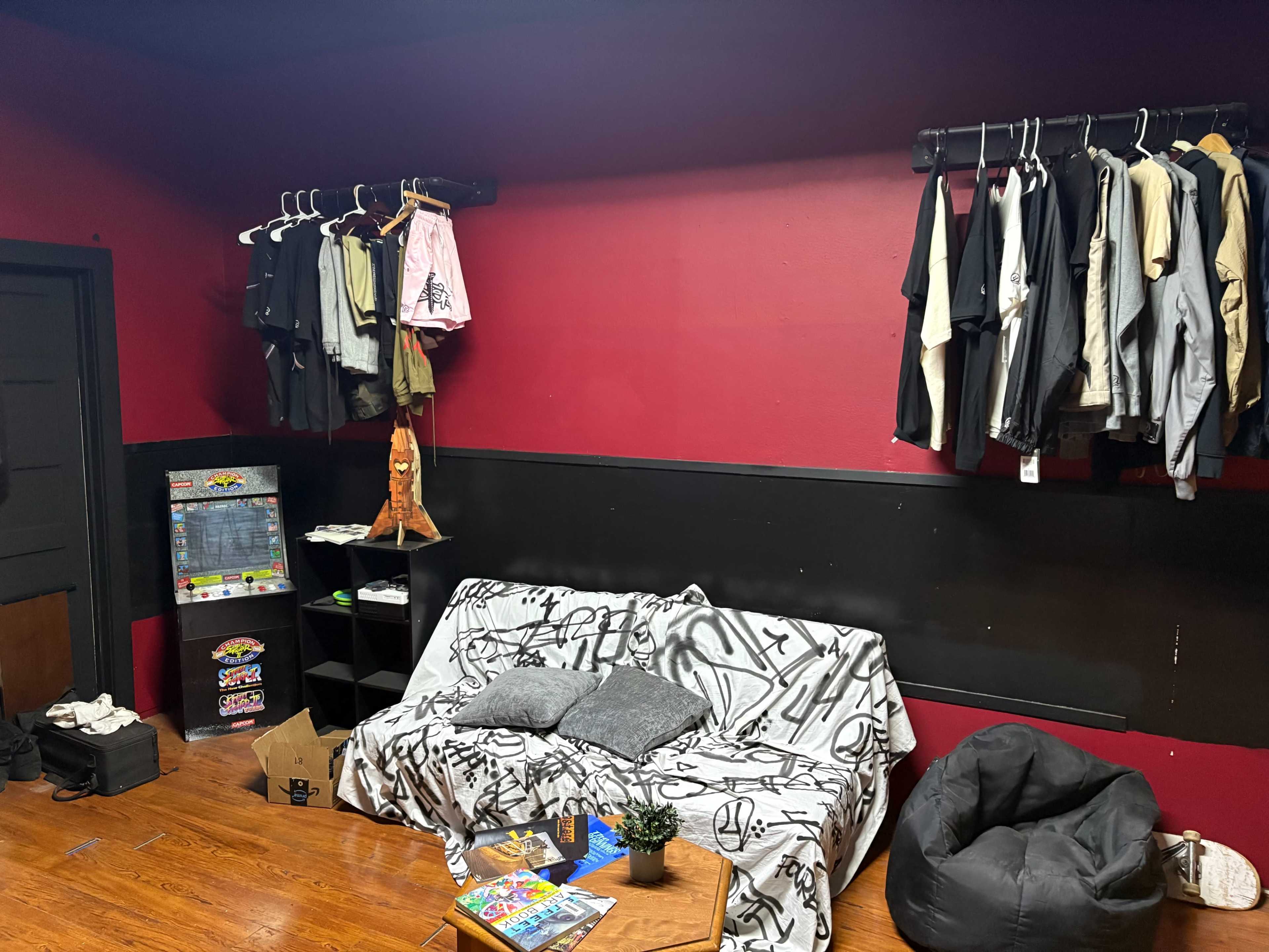 The image shows a small room with a couch covered by a white blanket, hangers with clothes on the wall, a bookshelf with various items, and a bean bag chair on the floor.