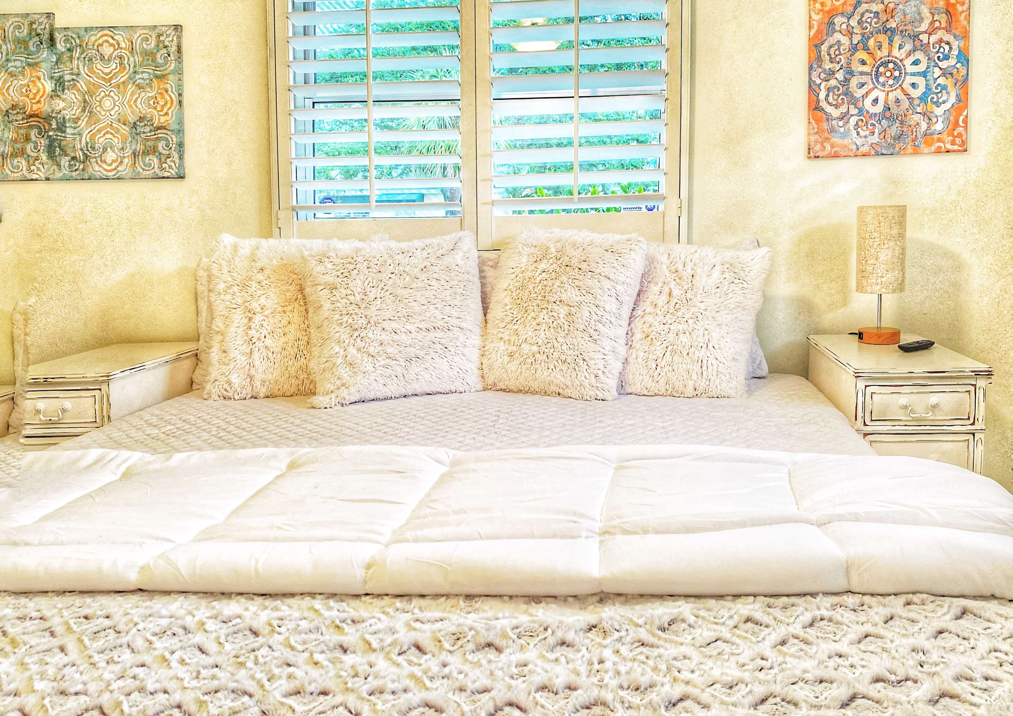 A neatly arranged bed with fluffy pillows and a white comforter is positioned against a wall with window shutters and decorative wall art.