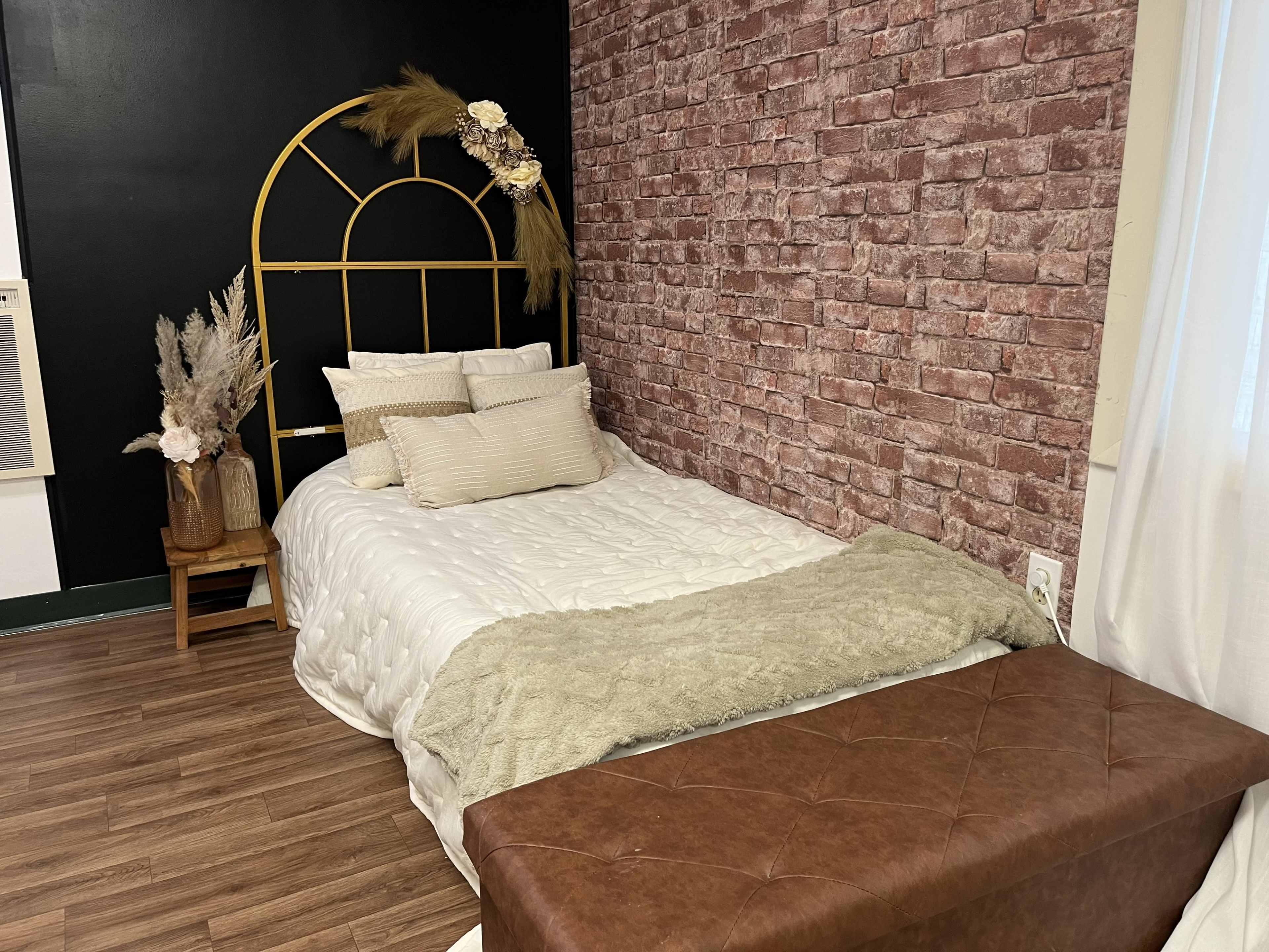 The image shows a cozy bedroom with a bed featuring a gold arched headboard, layered with white and beige bedding, against a brick-patterned wall.