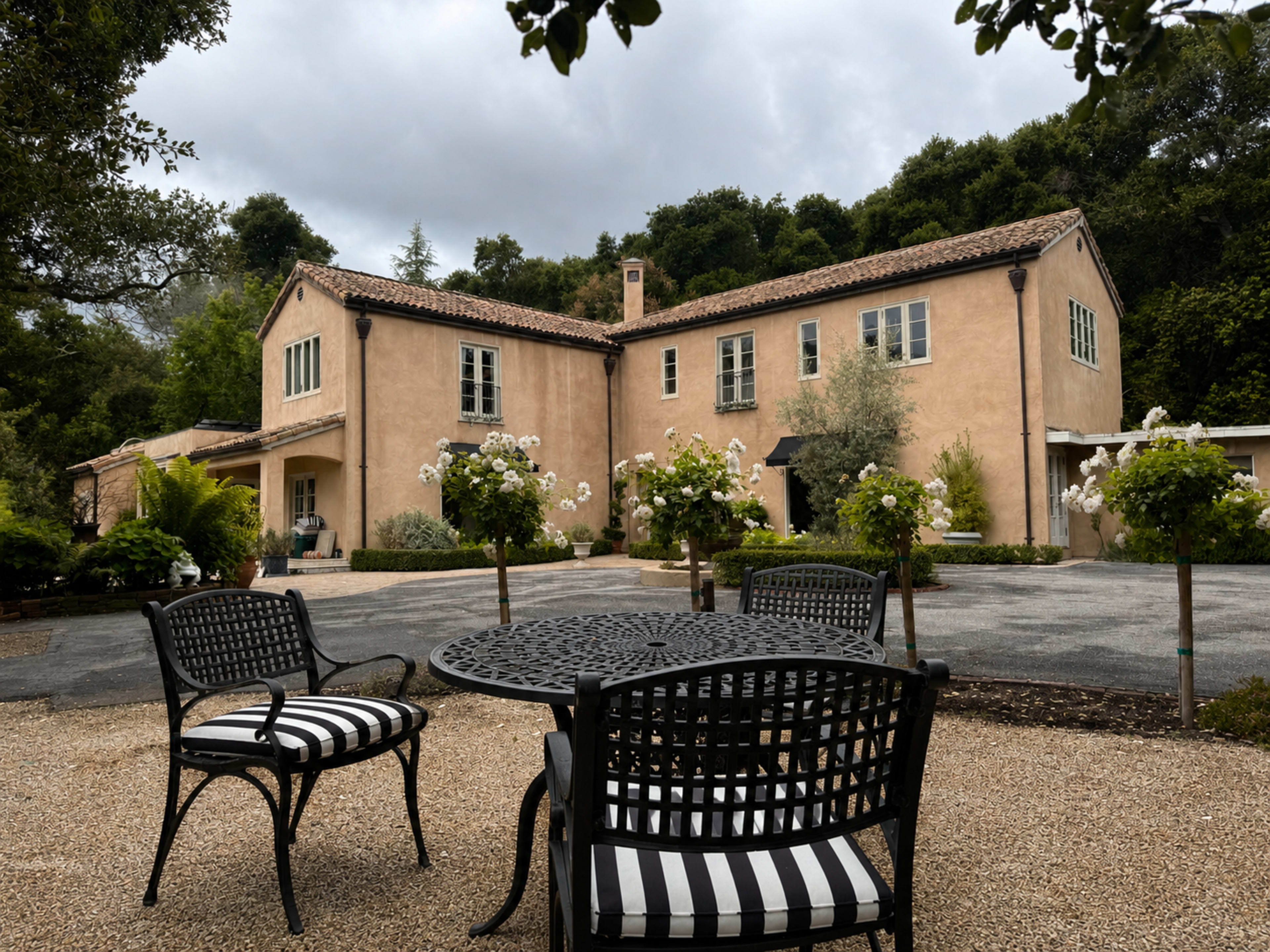 The image shows a two-story, stucco house surrounded by greenery, with a gravel driveway and black and white outdoor furniture in the foreground.