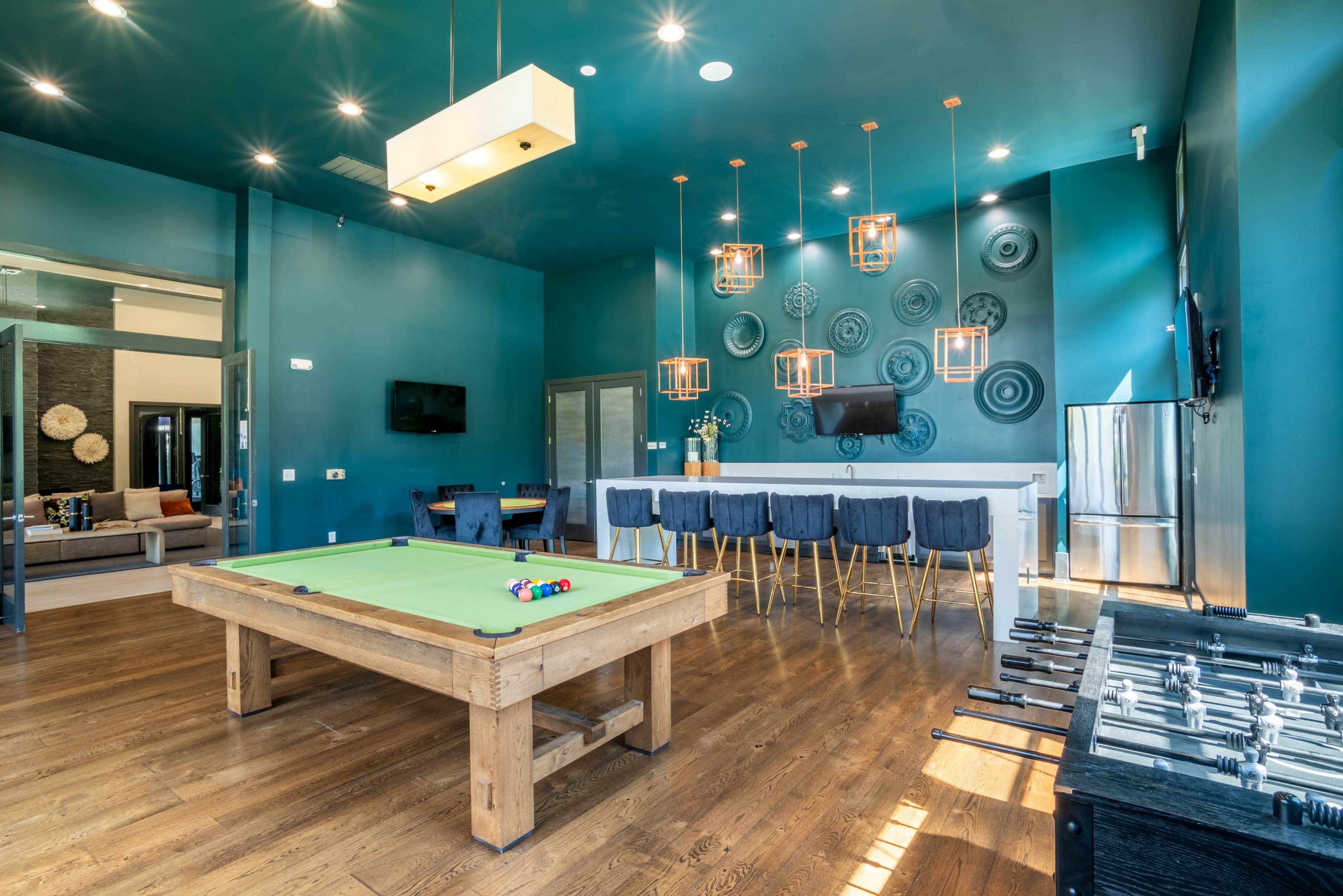 A game room featuring a pool table, a foosball table, a bar area with high stools, and various decorative elements on the walls.