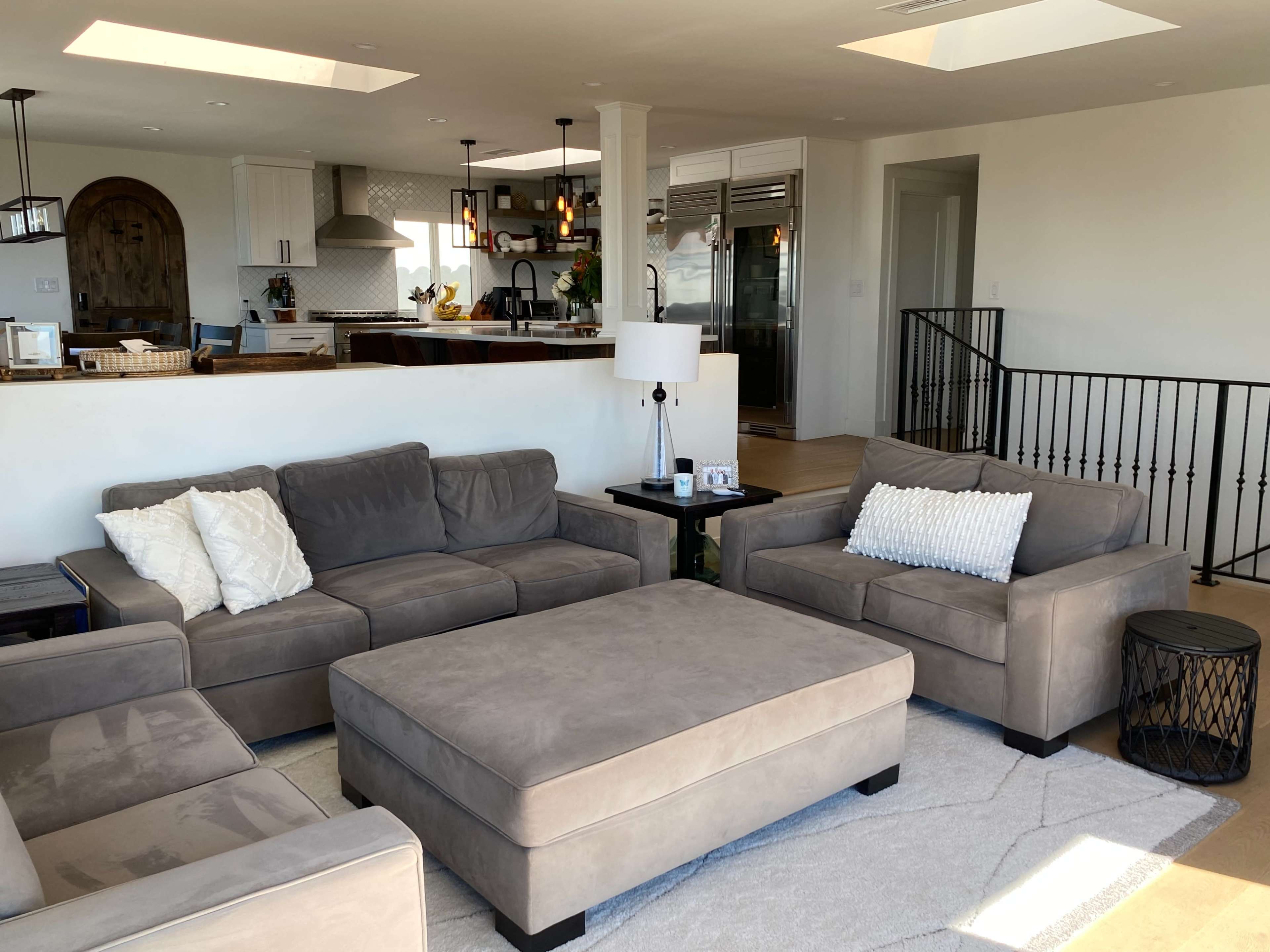 The living room features a gray sectional sofa and ottoman, with a lamp and decorative pillows, while an open kitchen is visible in the background.